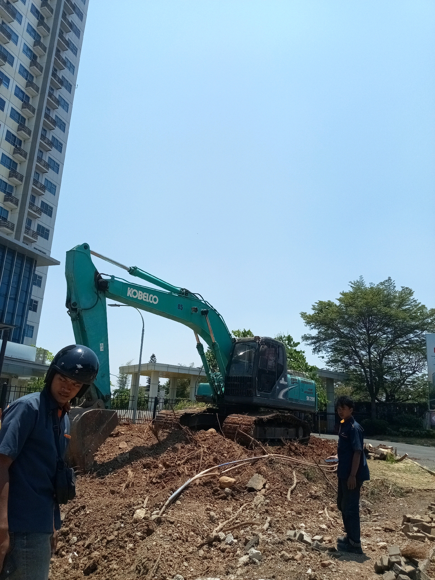 A construction site in Bekasi, Indonesia, is depicted under a clear blue sky. The scene features a large, green Kobelco excavator positioned on a mound of dirt and debris. Two individuals, presumably construction workers, are present. The person in the foreground, wearing a dark blue shirt, dark pants, and a black helmet, stands to the left, looking forward. To the right, another individual, also in a dark blue shirt and dark pants, is facing away from the camera. A tall, modern building with many windows and balconies is visible on the left side of the image, contrasting with the rustic construction environment. Trees and a street are also part of the background, indicating an urban or suburban setting within Bekasi.