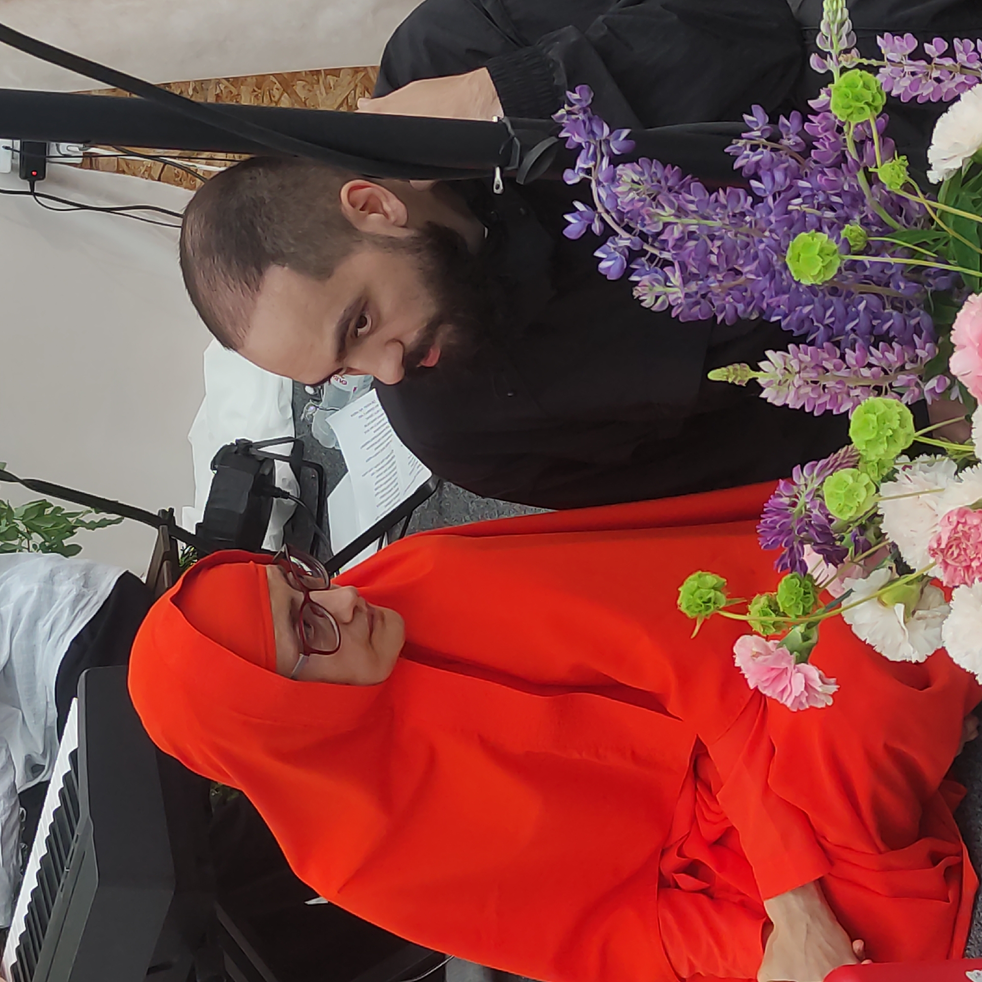 Two individuals are seated closely together, engaged in conversation. The person on the left wears a bright orange robe and glasses. The person on the right, with dark hair and a beard, wears a black jacket. In the foreground, a bouquet of flowers, including purple lupine, pink and white carnations, and green spherical blooms, is visible. Behind the individuals, a black electronic keyboard is partially visible, along with a metal stand and a white sheet. The background consists of a plain wall. The scene takes place indoors.