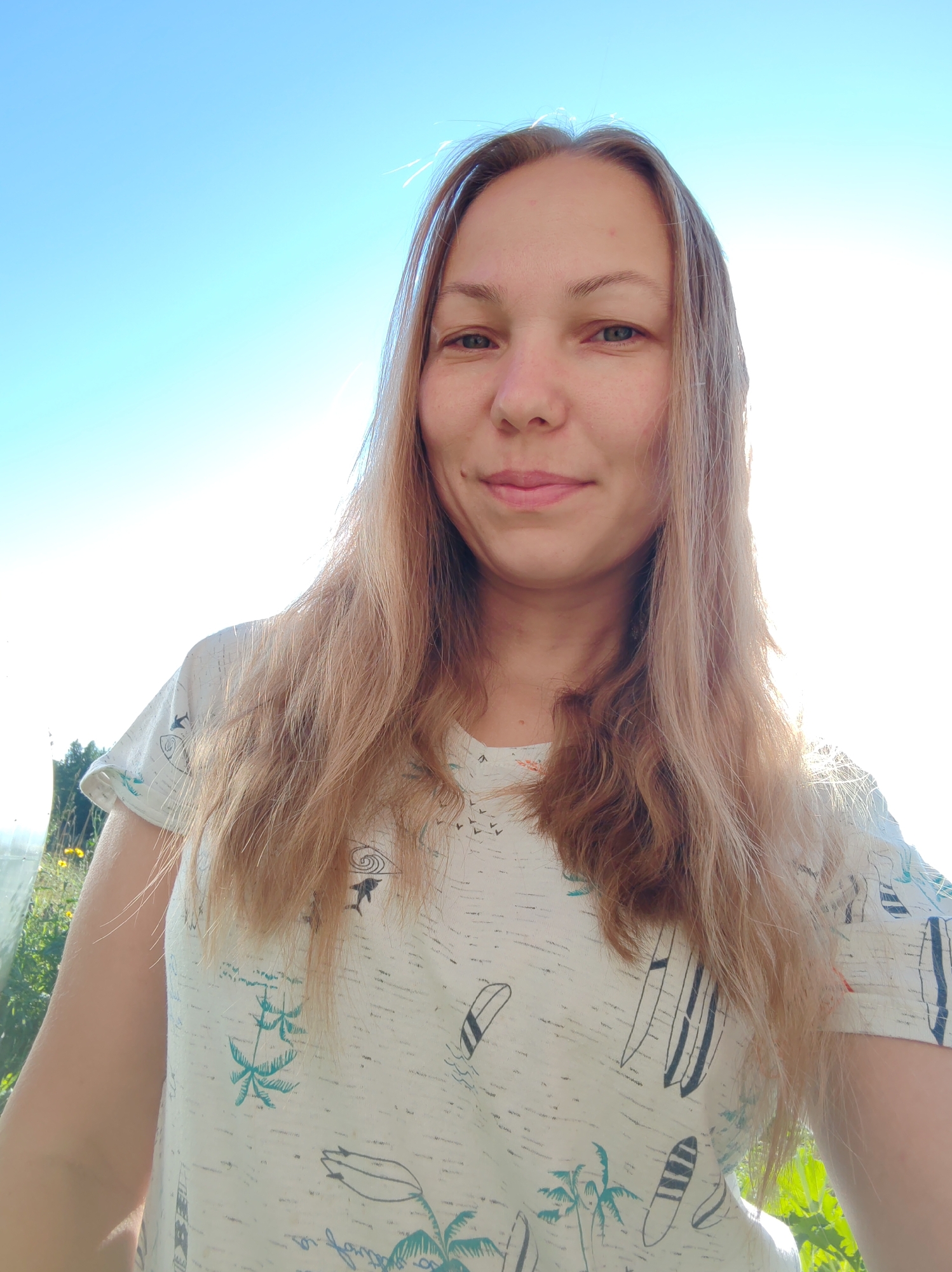 A young woman with long, blonde hair is pictured in a close-up shot against a bright blue sky. She is wearing a light-colored t-shirt with a pattern of palm trees and surfboards. The woman is looking directly at the camera with a slight smile. The sun is bright, creating some lens flare and blowing out the background in some areas. In the lower left and right corners, glimpses of green foliage and possibly a greenhouse structure are visible, suggesting an outdoor setting, potentially in or around Chukhlem, Russia.