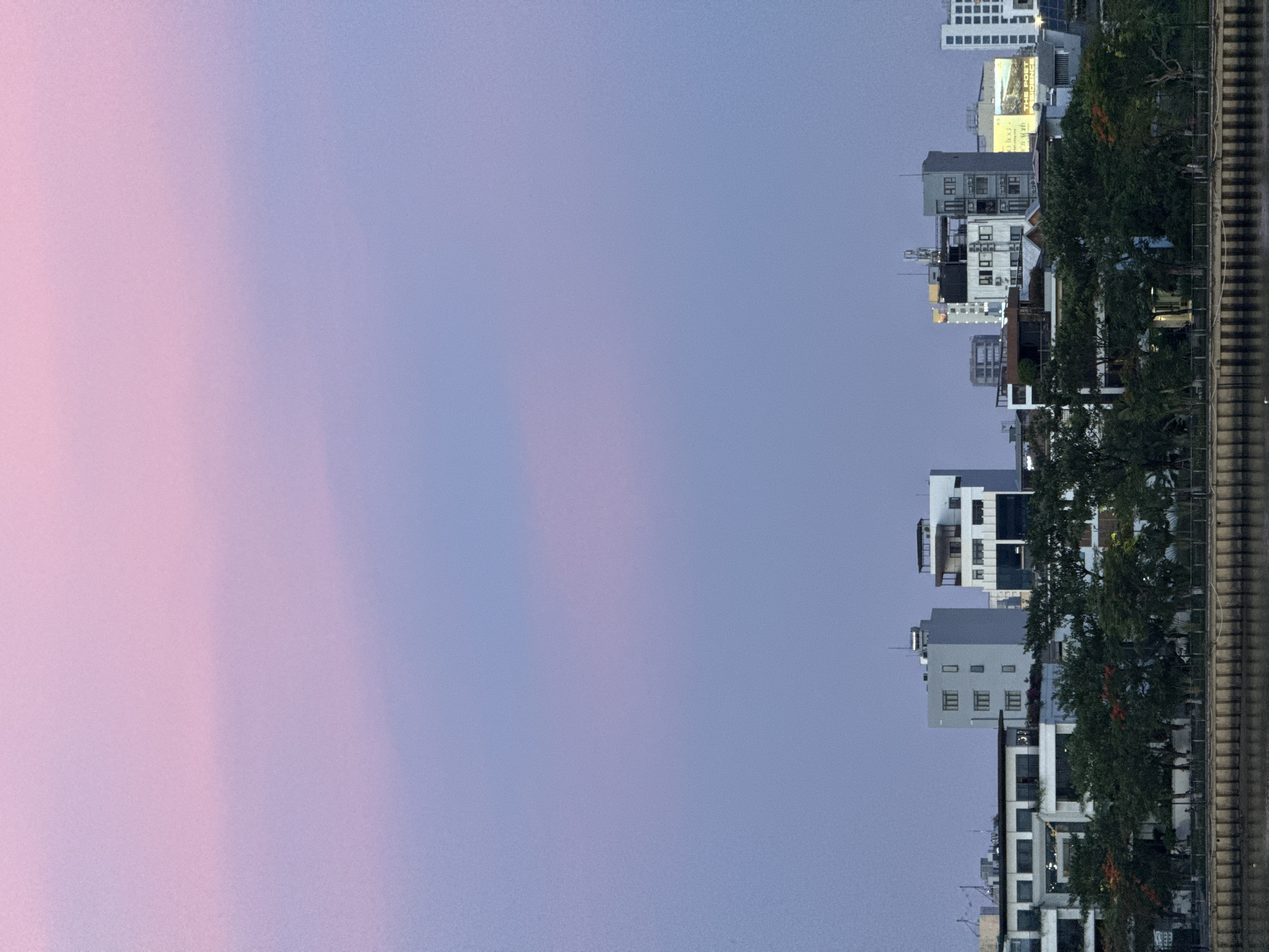 The image depicts a waterfront cityscape at dusk in Đà Nẵng, Vietnam, characterized by a gradient of soft pink and lavender hues in the sky above. In the foreground, a body of water, likely the Han River, is visible, bordered by a textured retaining wall. A line of lush green trees with some scattered red blossoms runs along the bank, separating the water from a row of modern buildings. These structures vary in height and architectural style, with some featuring balconies and large windows, and others exhibiting more minimalist designs. Several buildings are multi-story, with visible antennae and rooftop elements. The overall impression is of a tranquil urban evening scene.