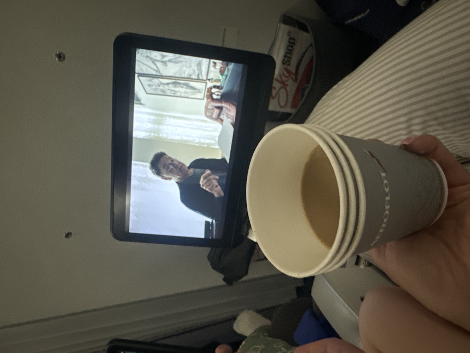 On an airplane, a passenger holds an Aeroflot paper cup filled with coffee in the foreground. In the background, an in-flight entertainment screen displays a man in a dark sweater, likely an actor in a movie. A Sky Shop advertisement is visible next to the screen. Striped fabric, possibly a blanket, is partially seen.