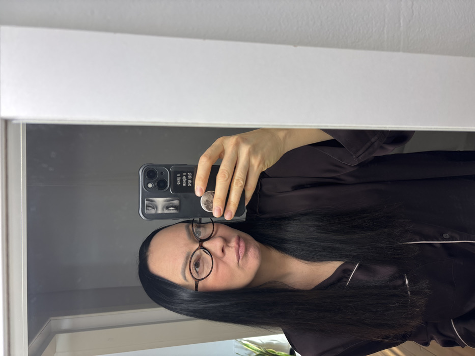 A woman with long black hair and round glasses is taking a selfie in front of a mirror. She's wearing a dark silky pajama set. Her phone case is black and has eye stickers and a pop socket. The background shows a white wall and part of a window with light filtering through. The image is reflected in the mirror.