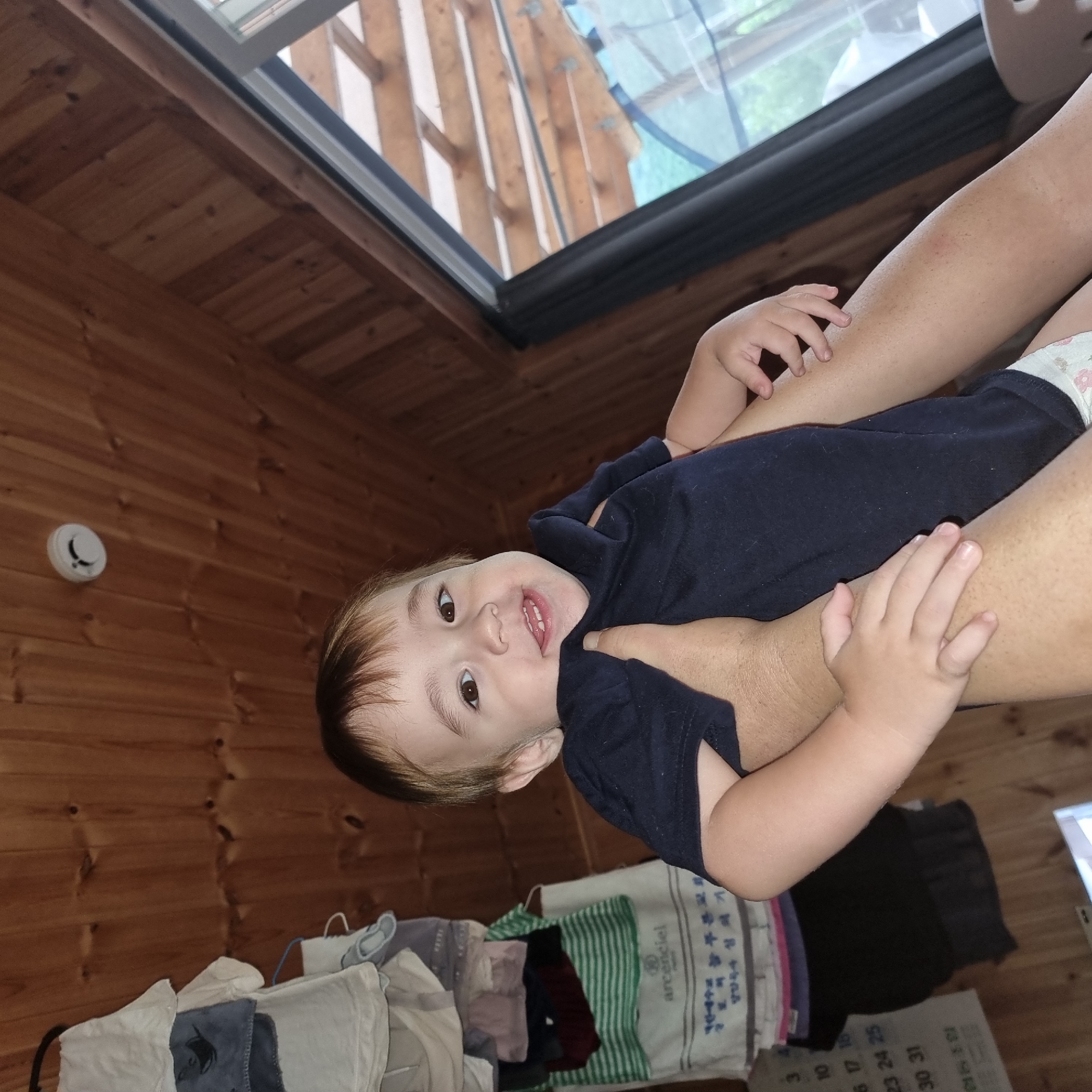 The image shows a young child being held by an adult. The child is wearing a dark blue shirt and a diaper with a floral pattern. The child has brown hair and brown eyes and appears to be smiling. The adult's arms are visible, supporting the child.
The scene appears to be indoors, with wooden paneling on the walls and floor. There is a skylight or window at the top of the image, showing a view of what looks like trees and sky, suggesting it is daytime. On the wall, there is a white circular object that resembles a smoke detector. In the foreground, there is a pile of folded laundry, including items with green and white stripes, and a white towel with text on it.
The overall mood seems to be casual and intimate, as the adult is holding the child close. The lighting suggests it is daytime. There is some visible text on a towel or fabric in the foreground which is partially obscured. The location is specified as Dunnae-myeon, South Korea.