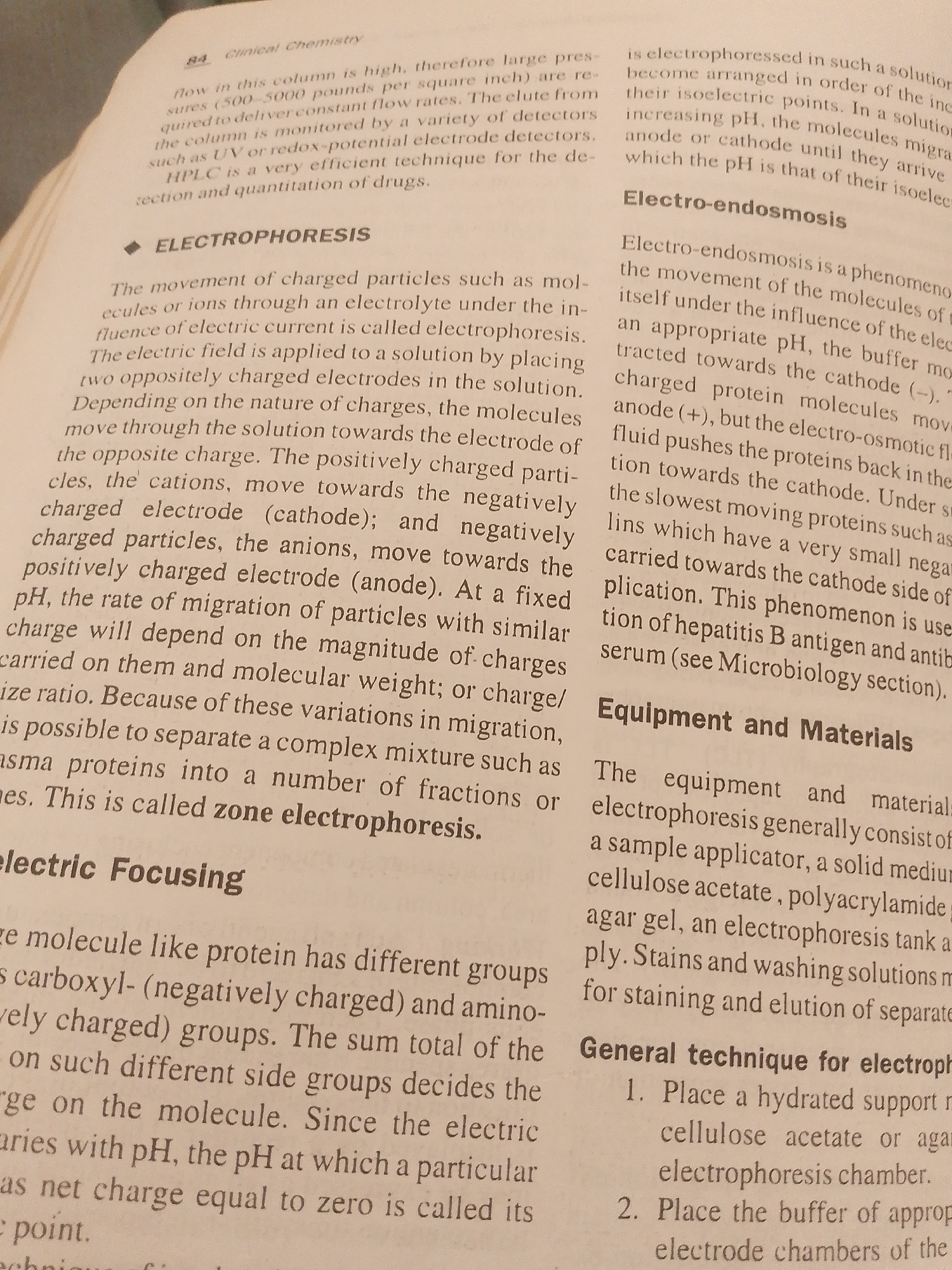The image is a close-up shot of an open book, showing two pages of text. The book appears to be a textbook on clinical chemistry, as indicated by the heading "Clinical Chemistry" at the top of the left page. The text discusses various electrophoresis techniques, including general electrophoresis, electro-endosmosis, and electric focusing, along with equipment and materials used. It also mentions HPLC as a drug detection and quantification technique. The pages are a light cream color and slightly textured, suggesting they are made of paper. There are no people, objects other than the book itself, or any indications of a specific scene, setting, time of day, or weather. The dominant content is the printed text, which is clear and legible in the provided crops.