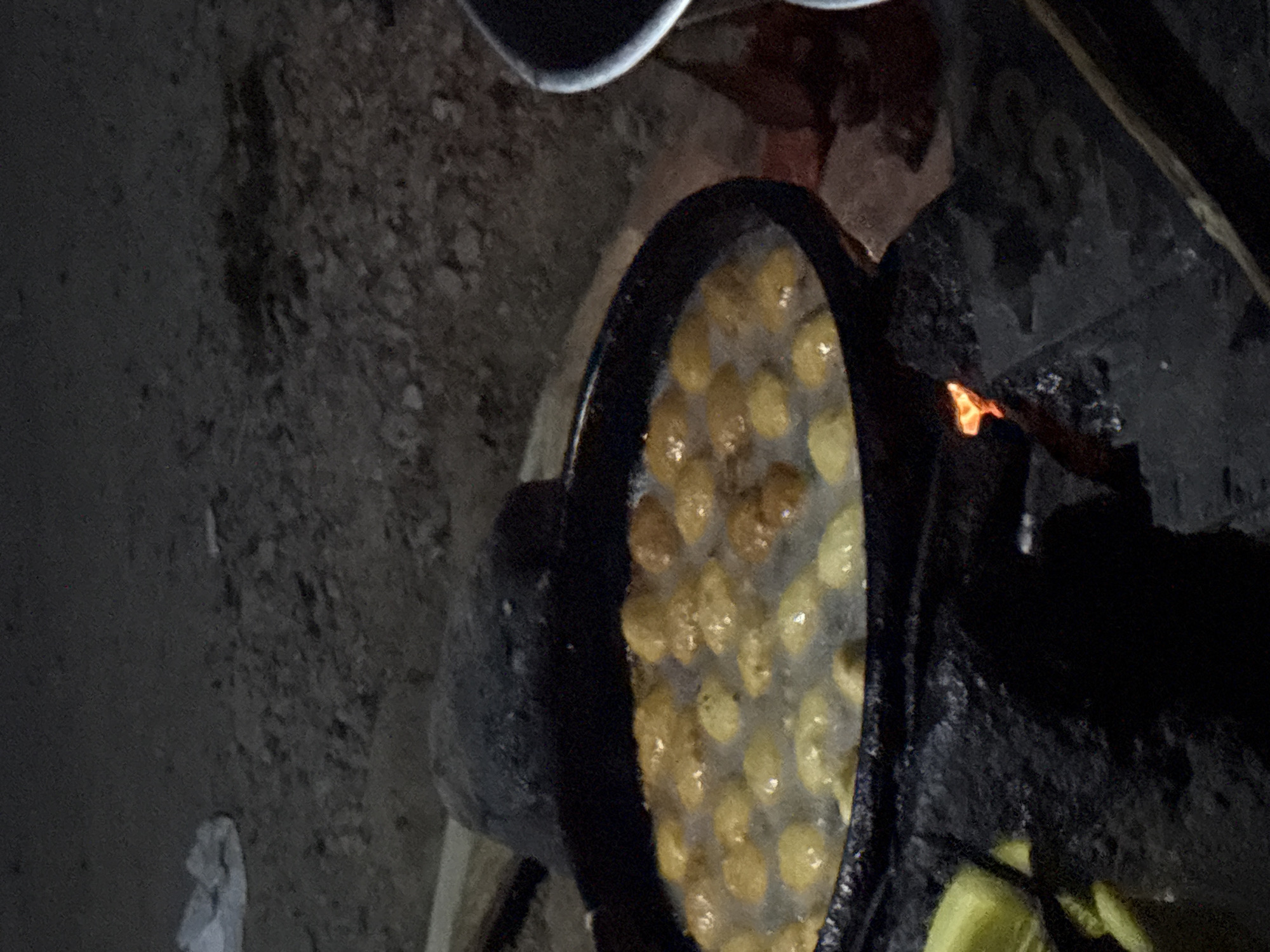 In the dim light of what appears to be nighttime, a large, black, cast-iron pot is positioned over a fire pit constructed with dark, rough stones. The pot is filled with what seems to be hot oil in which numerous round, golden-brown fritters are cooking. The fritters are submerged and bubbling, indicating they are actively being fried. Some of the fritters appear to have just been placed in the oil, while others are closer to being done. A large, dark, irregularly shaped object, possibly a cooking stone or a lid, is placed behind the pot. In the foreground to the left, partially visible green food items, possibly plantains, are present. The background is dark and out of focus, showing the rough texture of the ground, which is covered in small stones and dirt. A white piece of fabric or plastic is visible in the upper left corner of the image. The overall scene suggests an outdoor cooking setup in a rural or informal setting, potentially in the city of Angwan Sayawa, Nigeria, where traditional cooking methods over open fires are common.