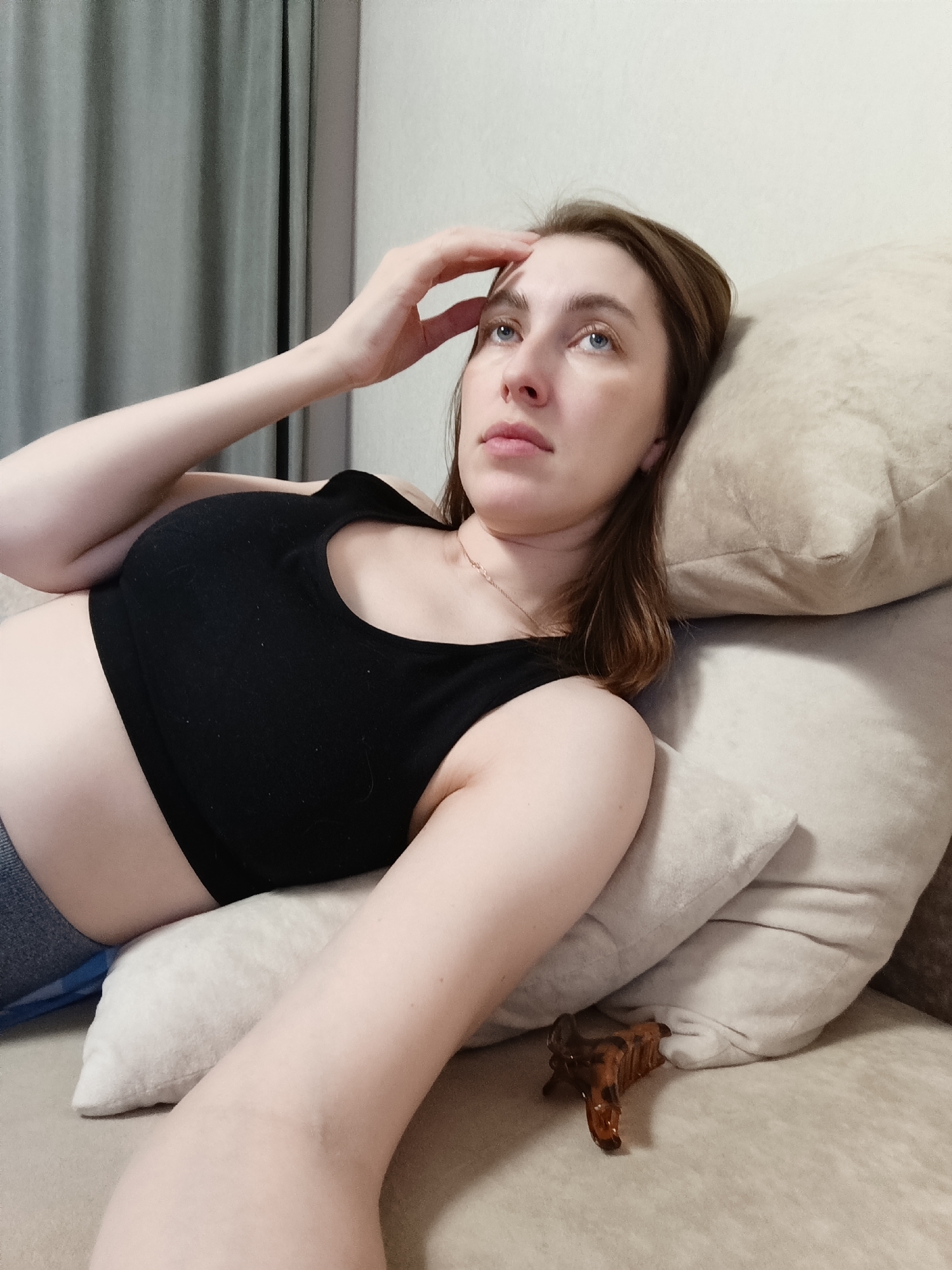 A fair-skinned young adult female lies on a tan couch, her head resting on two pillows. She wears a black sports bra and gray bottoms. Her right hand is placed on her forehead, and she gazes upward. A brown hair clip rests near the bottom of the pillows. A neutral-toned wall and curtain serve as the background. The location in the city of Chaikovsky, Russia, cannot be determined from the image alone.