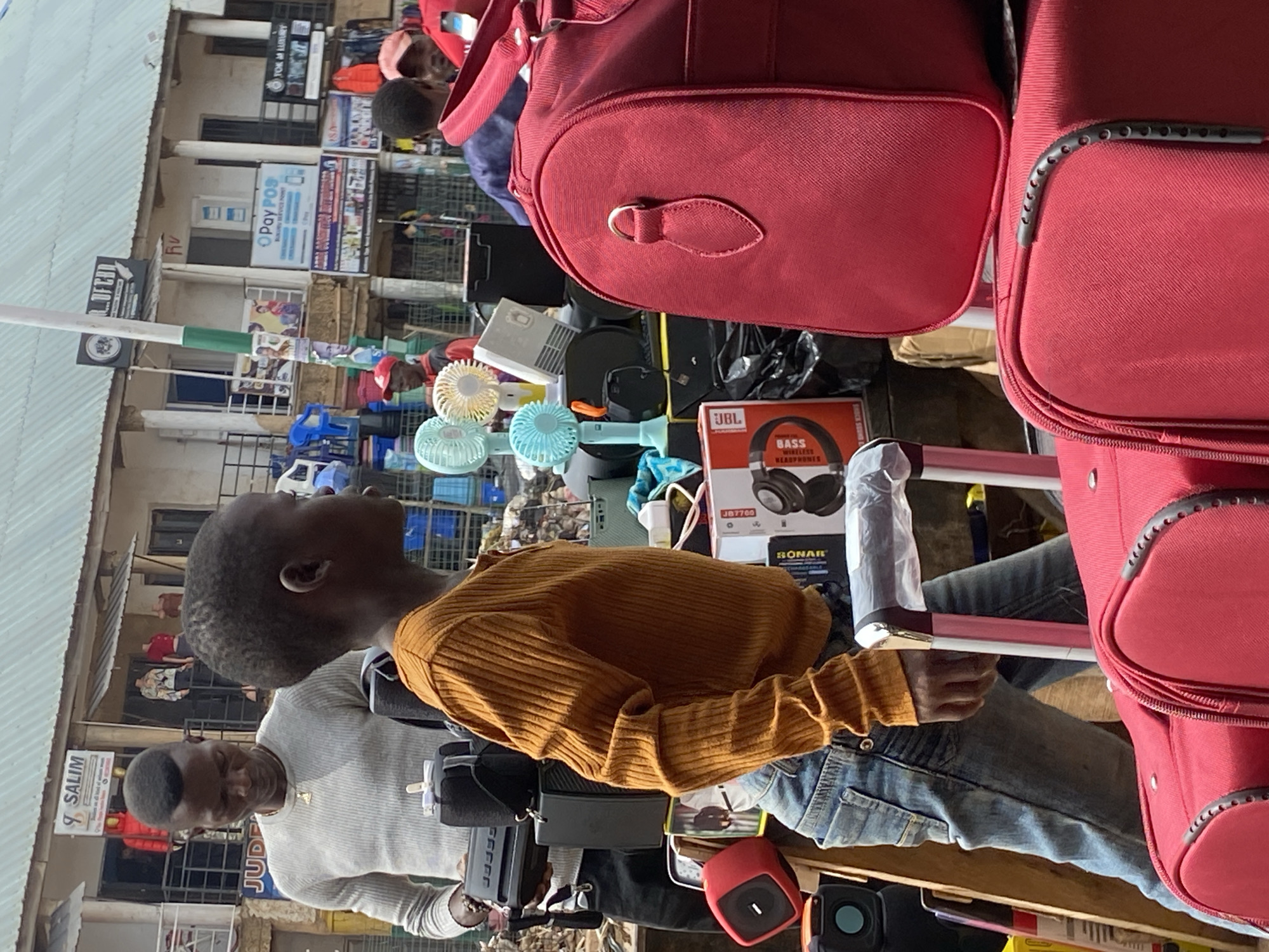 A busy outdoor market scene in Bukuru, Nigeria, is depicted. In the foreground, two red suitcases are stacked, one on top of the other. A boy, wearing an orange ribbed long-sleeved shirt and blue jeans, is partially visible, walking towards the right side of the frame. He appears to be looking ahead. Behind him, a man in a grey sweater is holding a black portable speaker. Several other objects are visible, including more speakers, small fans, and other market merchandise. In the background, shops with signs display various goods, with people milling about. The atmosphere suggests a lively commercial environment.