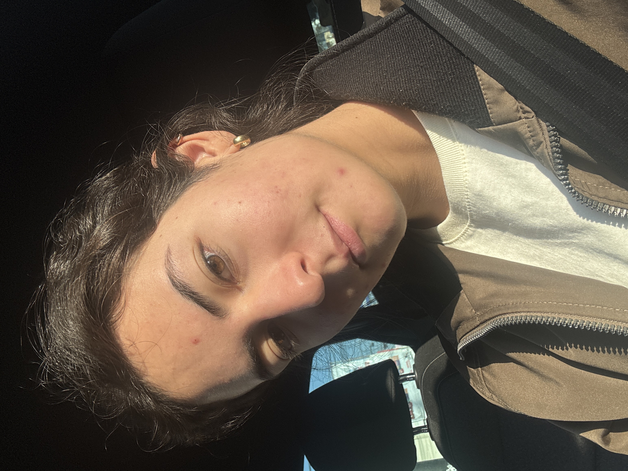 A young woman with dark curly hair, light skin, and brown eyes is in a car. She is wearing a light-colored shirt and a brown jacket with black stripes on the shoulders. She has a gold earring in her left ear and some blemishes on her face. The car's interior is mostly black. Through the car window, blurred buildings and a blue sky are visible, possibly indicating an urban environment in the city of Puebla, Mexico. The strong sunlight suggests it is daytime.