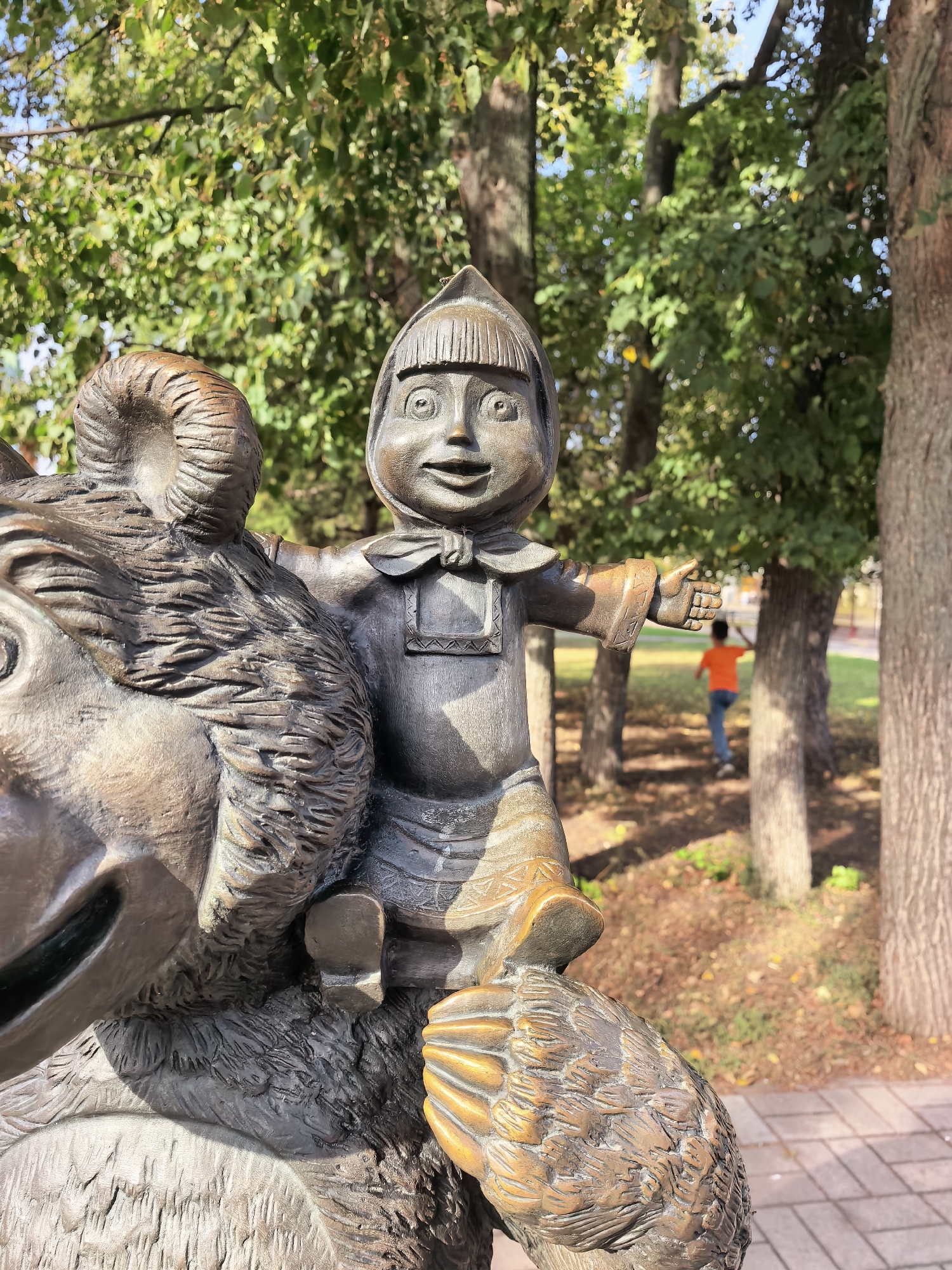 The image shows a bronze sculpture of a child riding a bear in a park setting. The child, with a cheerful expression, is wearing traditional clothing and a cap. The bear sculpture is detailed, with fur texture and a large head. In the background, a young boy in an orange shirt is running away from the camera. The environment appears to be a park with trees and fallen leaves on the ground, suggesting it might be autumn. The paving stones of a walkway are visible in the foreground. This sculpture is located in the city of Sterlitamak, Russia.