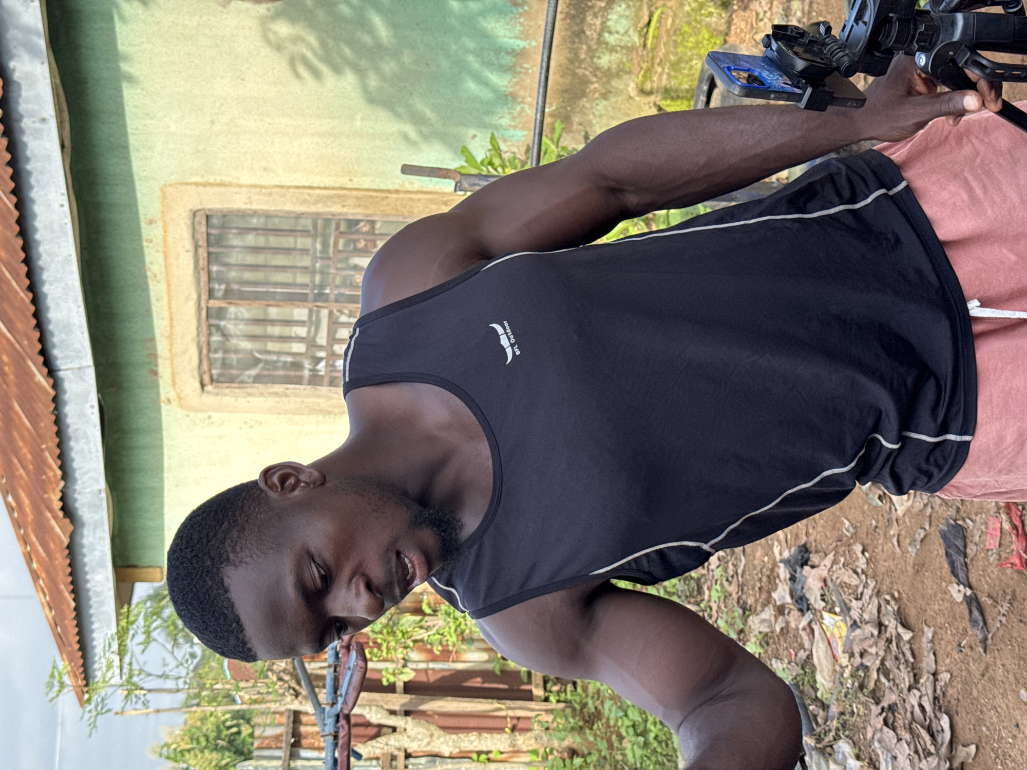 A Black man in a black tank top with white trim and pink shorts stands in front of a light green building with a rusty metal roof. The man is looking down to his left, with his mouth slightly open. His right arm is bent at the elbow, and his hand is resting on a black tripod with a phone mounted on it. The ground in front of the building is dirt and littered with trash, including plastic bags and dried leaves. Behind the man, a wooden fence is visible, and beyond that, green foliage. The building has a single window with metal bars. The scene appears to be outdoors in a rural or semi-rural setting, possibly in Akwanga, Nigeria.