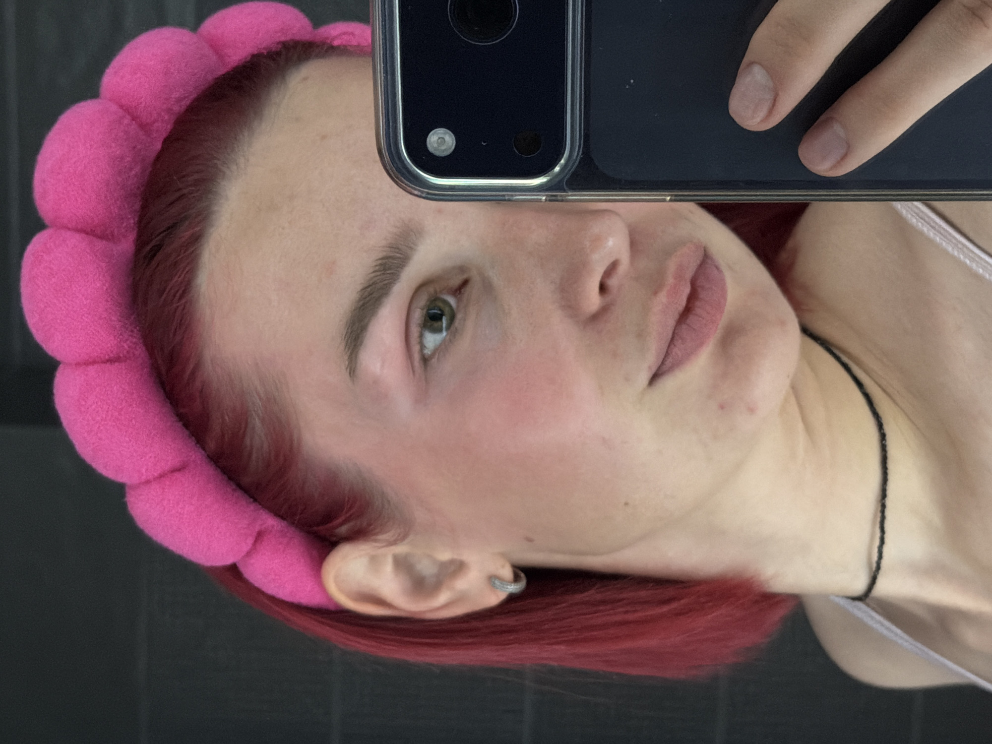 A young woman with bright red hair, wearing a puffy pink headband, takes a close-up selfie in what appears to be a bathroom. Her right eye, a striking green, is visible along with a portion of her nose and lips. A subtle sheen of makeup highlights her cheeks, and a thin black choker adorns her neck. She is holding a dark smartphone with a transparent case, its camera lens positioned to capture her reflection. The background is a dark, textured wall, suggestive of a tiled surface. The image does not provide any identifiable landmarks or elements that would suggest a specific location within the city of Chaikovsky, Russia.