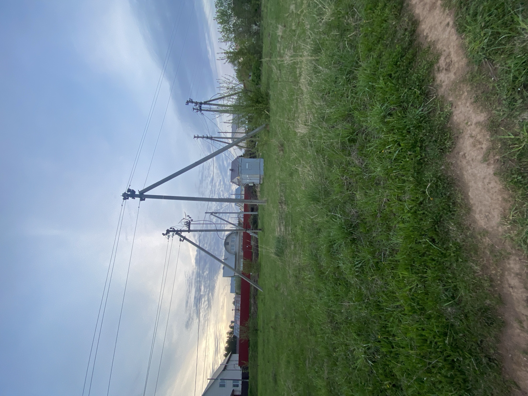 A low-angle, landscape-orientation shot displays a field with an unpaved path running alongside it, utility poles with power lines spanning across the upper-left portion of the frame, and a mix of trees, structures, and fences marking the boundary between the field and what appears to be a residential area. The location is Прикамский, Russia.
The sky occupies the upper-left portion of the frame, with a mix of clear blue and scattered clouds. The utility poles are positioned in a staggered line, with their support beams extending downwards towards the ground. Electrical wires are strung between the poles.
The field is primarily green, with variations in color indicating different types of vegetation. The unpaved path is visible to the right, its dirt surface contrasting with the green of the field.
Beyond the field, a red fence can be seen, along with hints of buildings. These structures suggest that the scene is at the edge of the town.