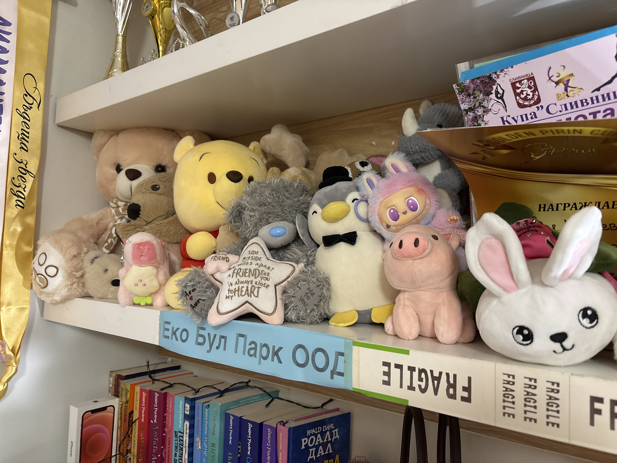 A shelf in what appears to be a child's room or play area is filled with stuffed animals, trophies, and books. The stuffed animals include various bears, a penguin, a pink pig, and a white bunny. Trophies are on the shelf above. The books on the lower shelf have titles in Bulgarian, and there is an iPhone box to the left. A sign reading "Еко Бул Парк ООД" is visible, indicating this may be a business or organization. A ribbon with Bulgarian text hangs on the left side. A sign with the word "FRAGILE" is repeated along the edge of the shelf. Another sign with text in Bulgarian "Купа Сливница" is visible in the upper right. The shelf's location is Svilvnitsa, Bulgaria. The city of Sofia, Bulgaria, is not directly identified in this image.