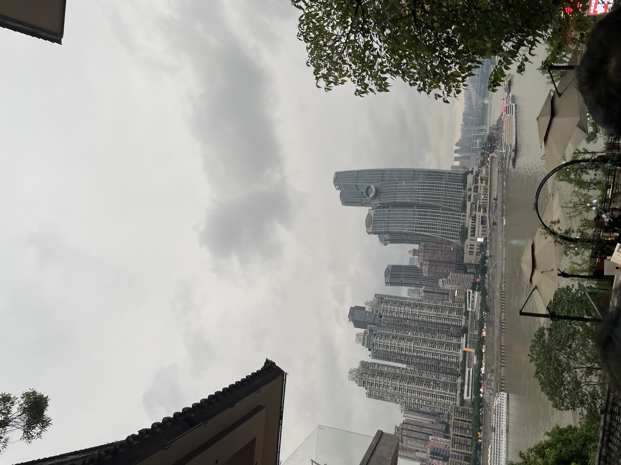 This image captures a view of the Chongqing Yangtze River Cableway in Nan'an District, China. The scene is dominated by a cloudy sky and a sprawling cityscape with numerous high-rise buildings. In the foreground, a traditional Chinese-style building with a dark tiled roof is partially visible on the left. A large river, likely the Yangtze River, flows through the center of the frame, with a white passenger boat navigating its waters. Several other boats are also present on the river. The cityscape features a cluster of modern skyscrapers, including one particularly distinctive building with a curved facade and a circular structure near its top. Trees and foliage are scattered throughout the scene, with a large tree on the right side of the image. The overall atmosphere is overcast, suggesting a cloudy day. The view appears to be from a vantage point overlooking the river and the opposite bank of the city.