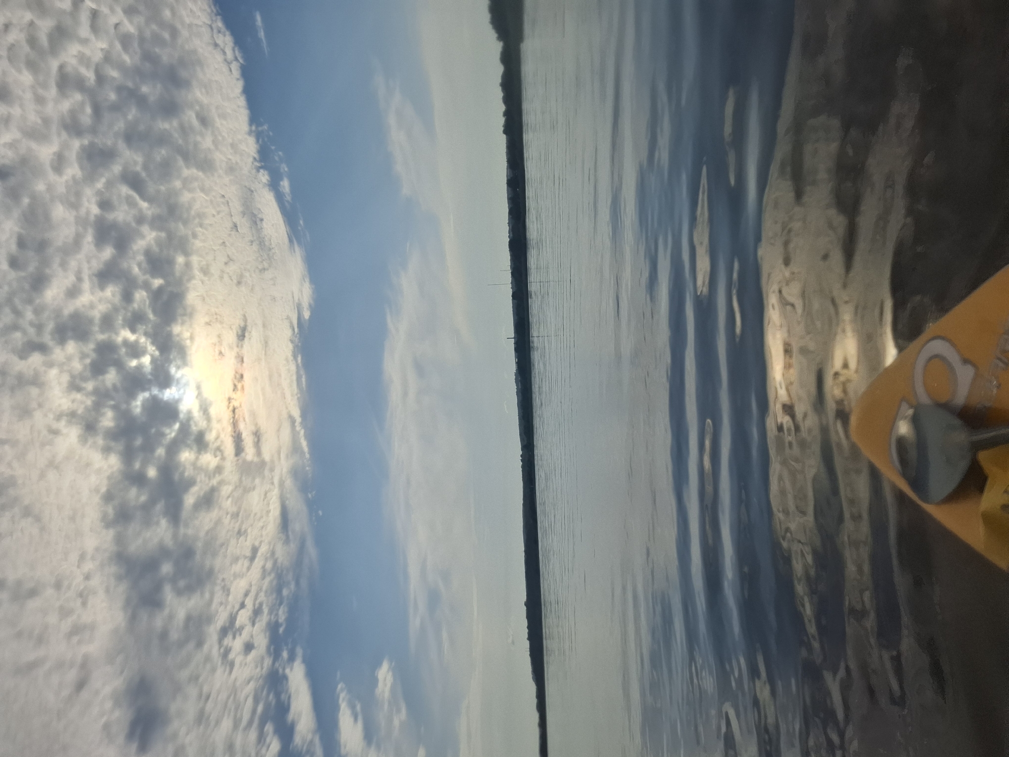 The image shows a wide body of water, likely a river or lake, viewed from a kayak. The water is calm with gentle ripples reflecting the sky. The sky is a bright blue with scattered white and grey clouds, and the sun is partially obscured by clouds, casting rays of light. In the distance, a tree-lined horizon is visible, with some faint structures that could be buildings or towers. The front of a yellow kayak is visible in the foreground, with a metal oar resting on it. The scene appears to be set in Svobodny, Russia, during daylight hours, possibly early morning or late afternoon given the lighting.