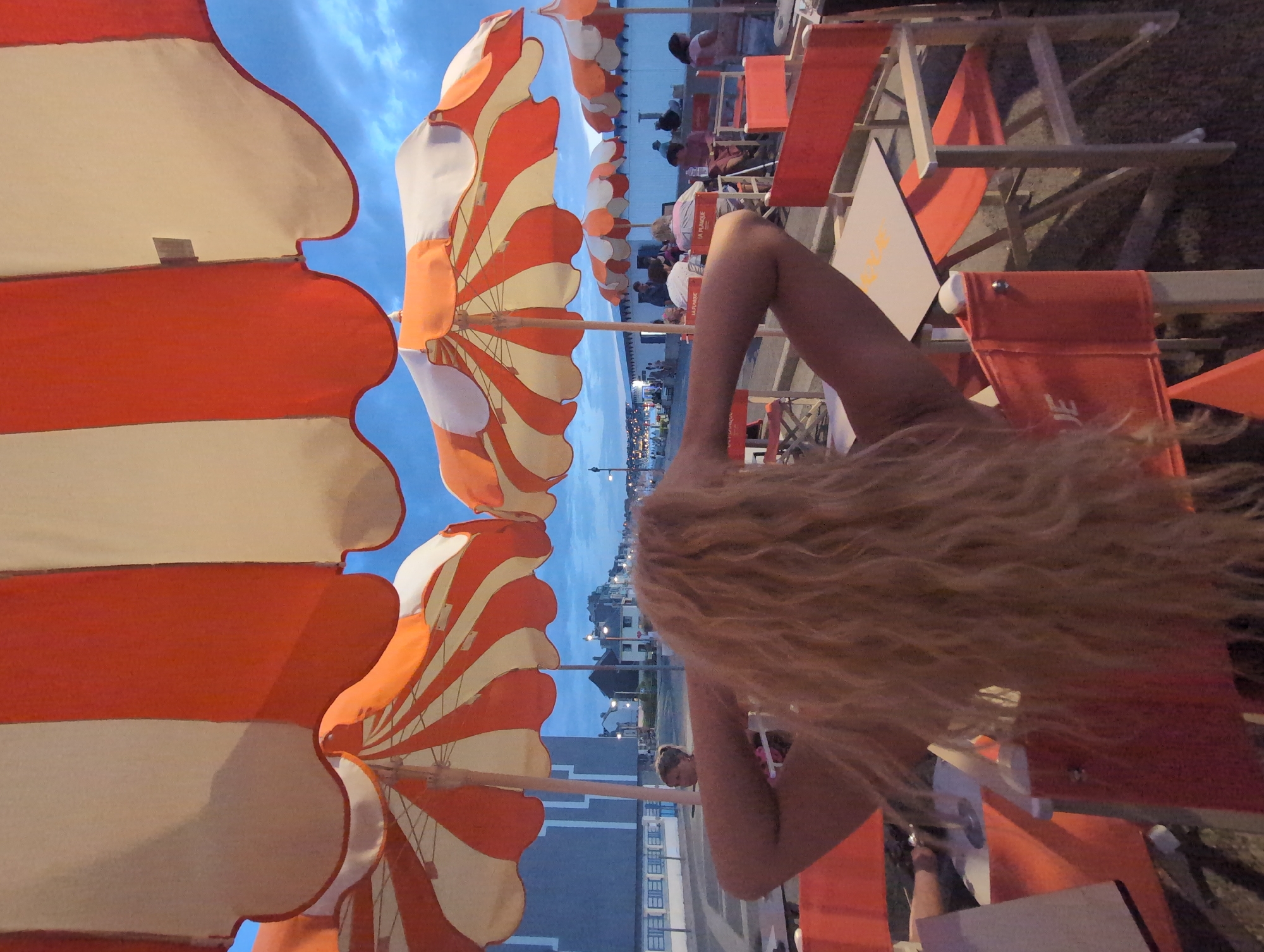 The image captures an outdoor dining scene in Mers-les-Bains, France, with a focus on a person in the foreground with blonde, curly hair, facing away from the camera and looking towards the sea. The person is sitting at a table with their arms behind their head, under a large, orange and white striped umbrella. Several other orange and white striped umbrellas are visible, some larger than others, casting shadows. Orange chairs and tables are set up on a paved area. In the background, a row of buildings with windows is visible along a walkway, suggesting a seaside promenade. Further in the distance, people can be seen sitting at tables and walking. The sky is a dusky blue, indicating it is either early morning or late evening. The overall ambiance is that of a relaxed, beachfront dining establishment.