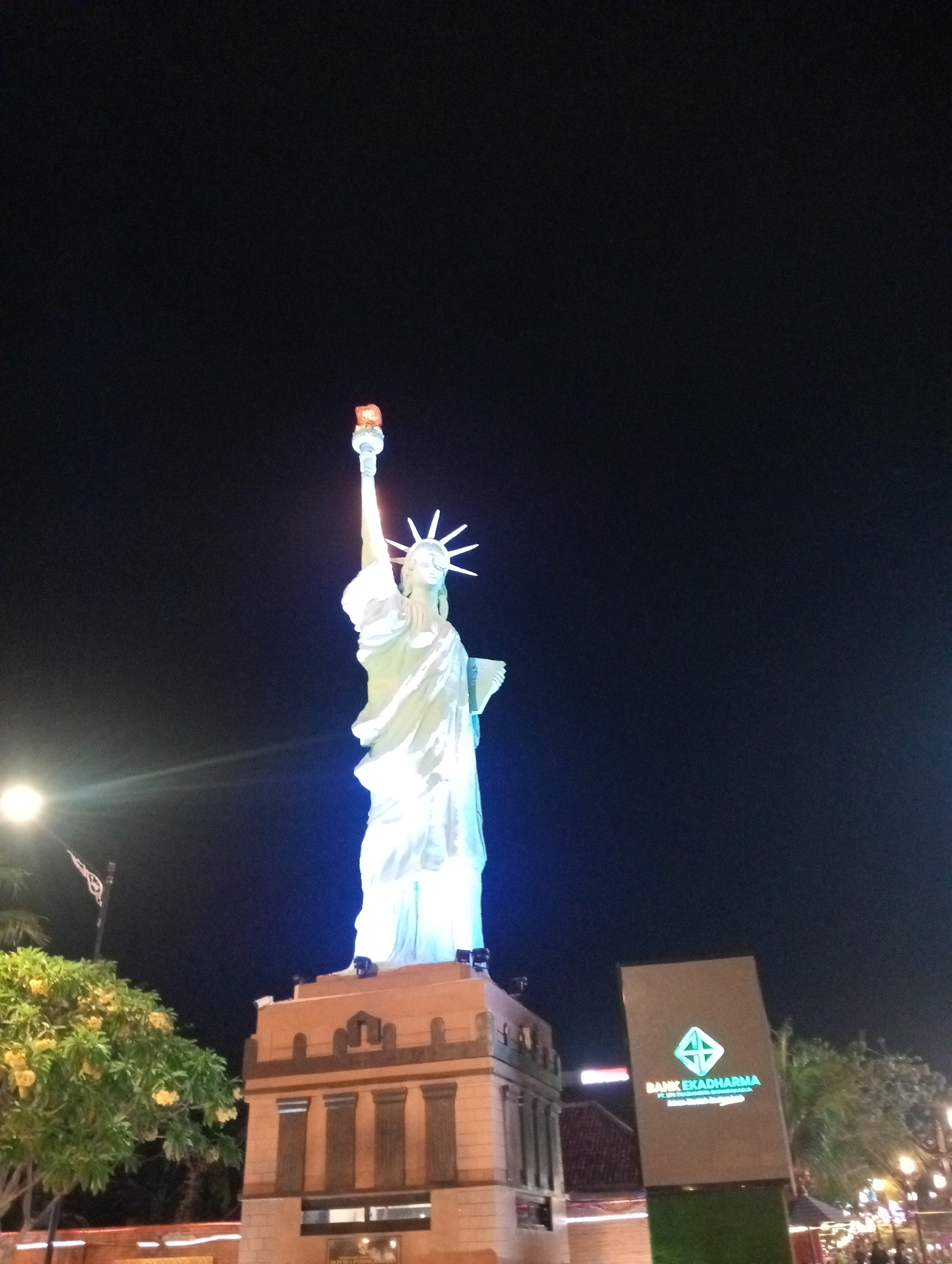 The image captures a nighttime view of a replica of the Statue of Liberty, illuminated with white and blue lights, standing on a pedestal in Madiun, Indonesia. The statue is depicted from a low angle, with its torch raised high. To the left of the statue, a bright streetlight casts a glare. In the foreground, a tree with yellow flowers is partially visible. To the right, a sign with the "BANK EKADHARMA" logo is illuminated. The background consists of a dark night sky and the lights of what appears to be a commercial area, including other illuminated signs and building lights. The scene suggests an outdoor public space, likely a tourist attraction or a commercial district in Madiun.
