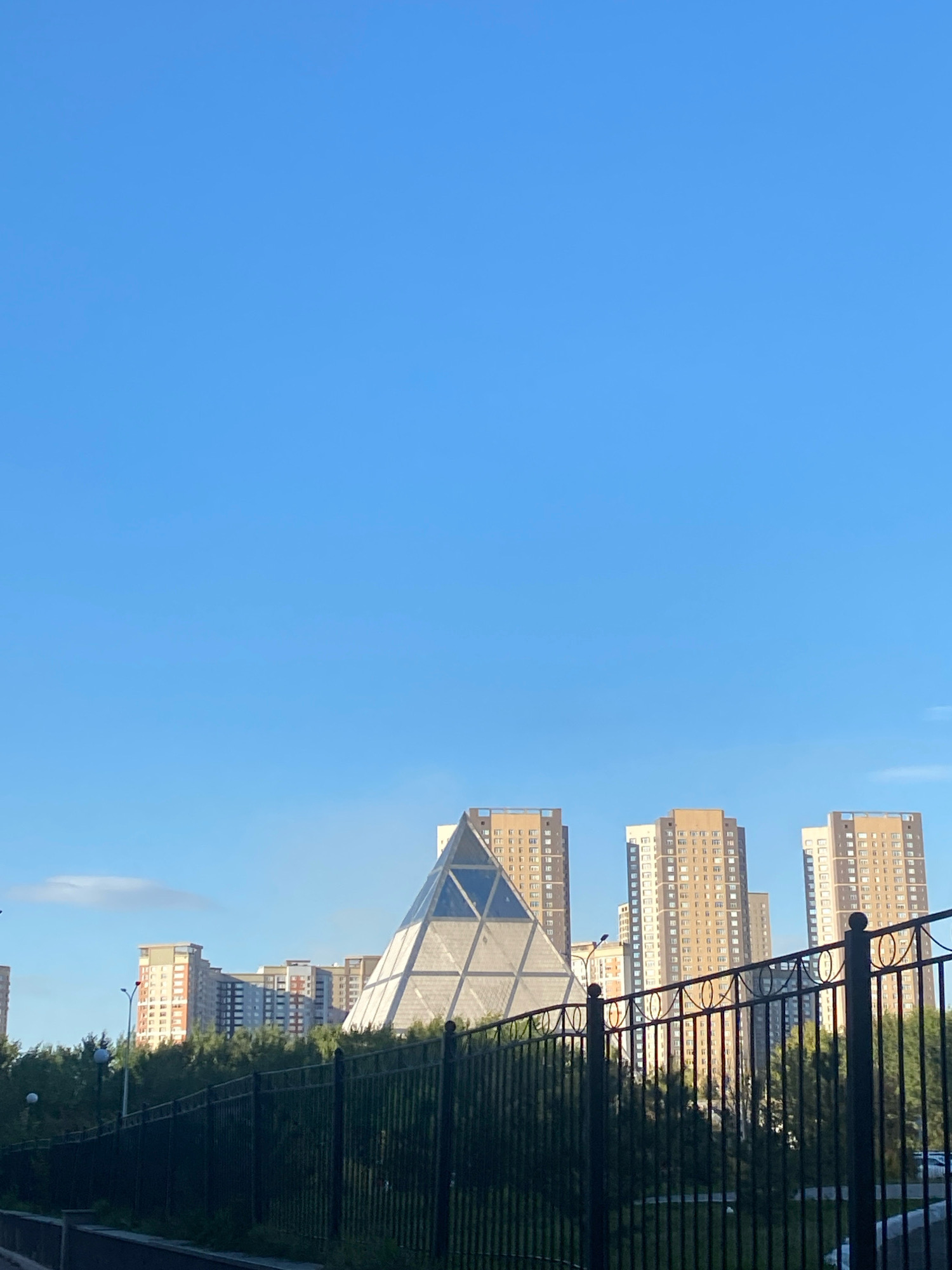 This image captures a panoramic view of the city of Astana, Kazakhstan, featuring a prominent pyramidal structure known as the Palace of Peace and Reconciliation. The sky is a clear, bright blue, taking up the majority of the frame. Below the sky, the pyramidal building stands out with its geometric design, illuminated by natural light. Surrounding the pyramid are several tall, modern residential buildings with numerous windows, characteristic of urban architecture. In the foreground, a black metal fence runs horizontally across the image, partially obscuring the view of lush green trees that stand between the fence and the buildings. The fence has a repeating pattern of circular shapes and vertical bars, suggesting a park or public area. The overall scene depicts an urban landscape under a bright, sunny sky.