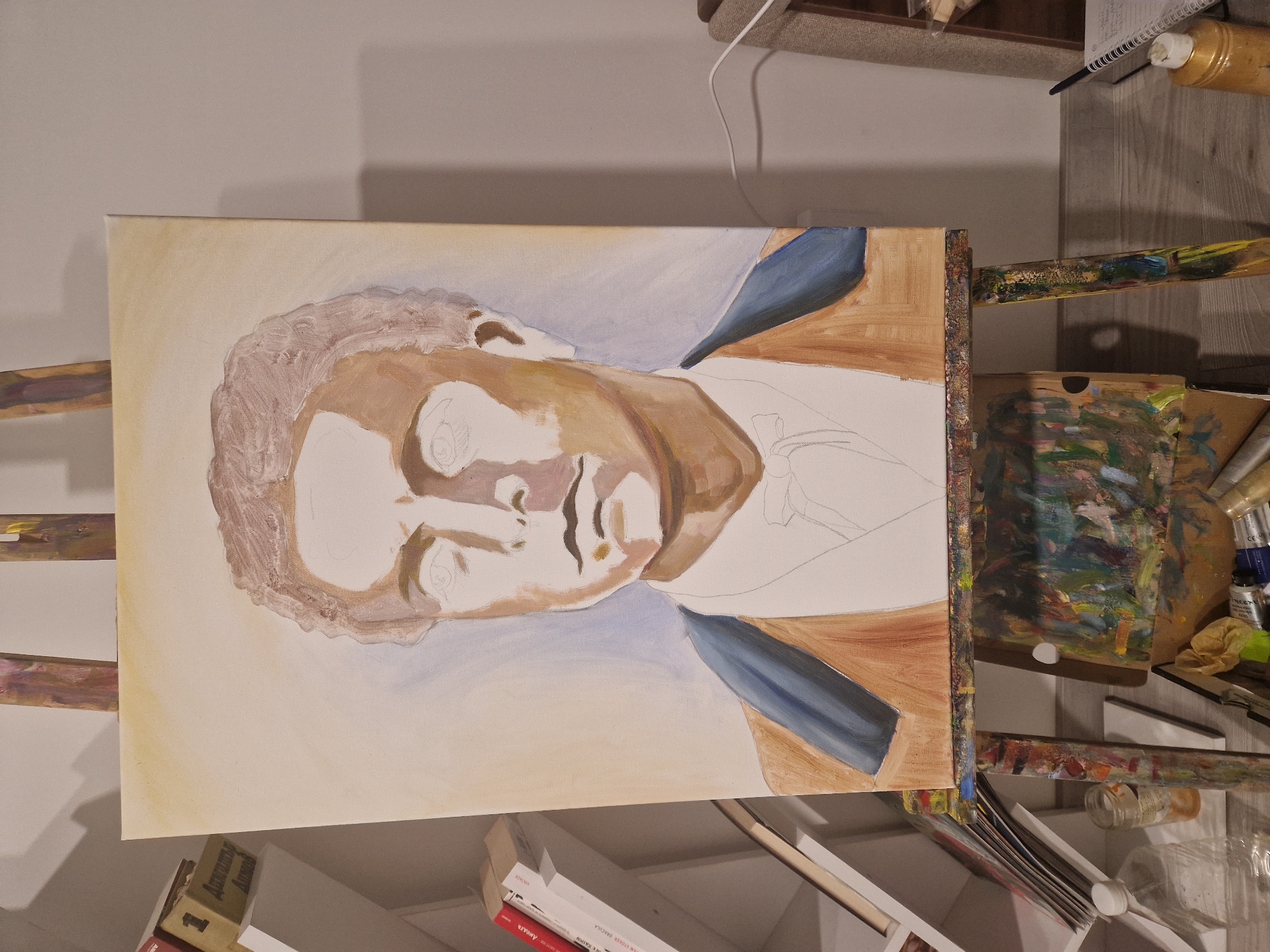 The image shows an unfinished portrait painting on a canvas mounted on an easel. The subject appears to be a male figure with partially rendered hair and face in varying shades of brown. His coat is indicated with broad strokes of brown and blue. The background has light yellow and blue hues. The easel is surrounded by art supplies, including paint tubes, brushes, and a palette covered in mixed paints. The scene is indoors, with a desk or table partially visible in the background, holding additional art supplies and books.