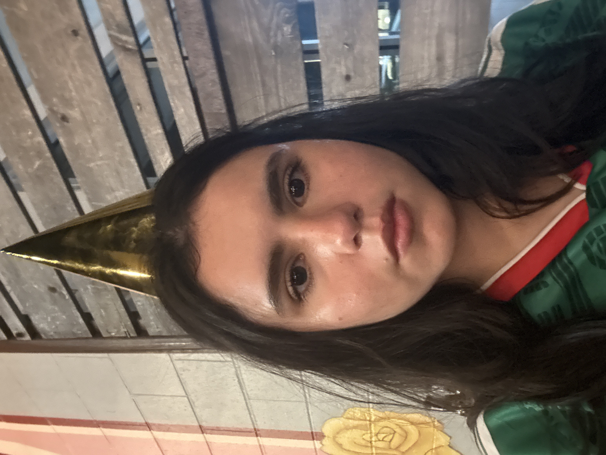 A close-up selfie shows a young woman wearing a gold party hat and a green and white striped shirt. She has long, dark hair and is looking directly at the camera with a neutral expression. The background consists of a light-colored brick wall with a floral design on the left, and a wooden slatted wall on the right. The lighting is soft and natural. The location is unspecified.