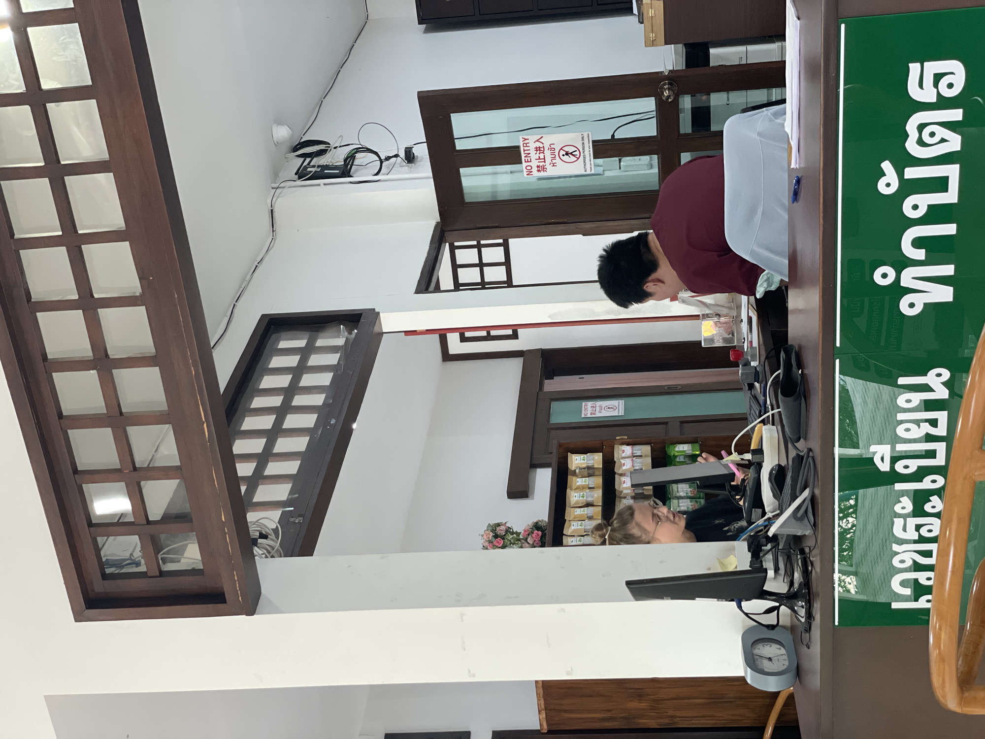 The image captures an indoor scene, likely a reception or administrative counter, in what appears to be an office or clinic within Chiang Mai City Municipality, Thailand. The dominant colors are white for the walls and ceiling, and dark brown for wooden architectural elements and furniture.
Two individuals are present, seemingly working at a counter. One person, with dark hair and wearing a burgundy shirt, is seen from behind, leaning over a dark wooden counter, focused on something below their line of sight. To the right, another person with light-colored hair and glasses is partially visible behind a different section of the counter, also engaged with tasks on the desk, possibly using a computer.
The counter features several items including computer monitors, keyboards, and a small grey analog clock. A prominent green sign with white Thai script, "เวชระเบียน ทำบัตร," is affixed to the counter, translating to "Medical Records / Make Card," which indicates the nature of the facility. Overhead and behind the counter, a tangle of wires, network equipment, and a security camera are visible.
A dark wooden door with a glass panel displays a "NO ENTRY" sign in English, Chinese ("禁止进入"), and Thai ("ห้ามเข้า"), with additional text "AUTHORISED PERSON ONLY" below it. Through another opening or doorway, shelves stocked with various packaged items and a small bouquet of artificial pink roses can be seen. The room features decorative dark wooden lattice structures, possibly part of partitions or window frames, adding to the interior design. The time of day is not discernible due to indoor lighting.