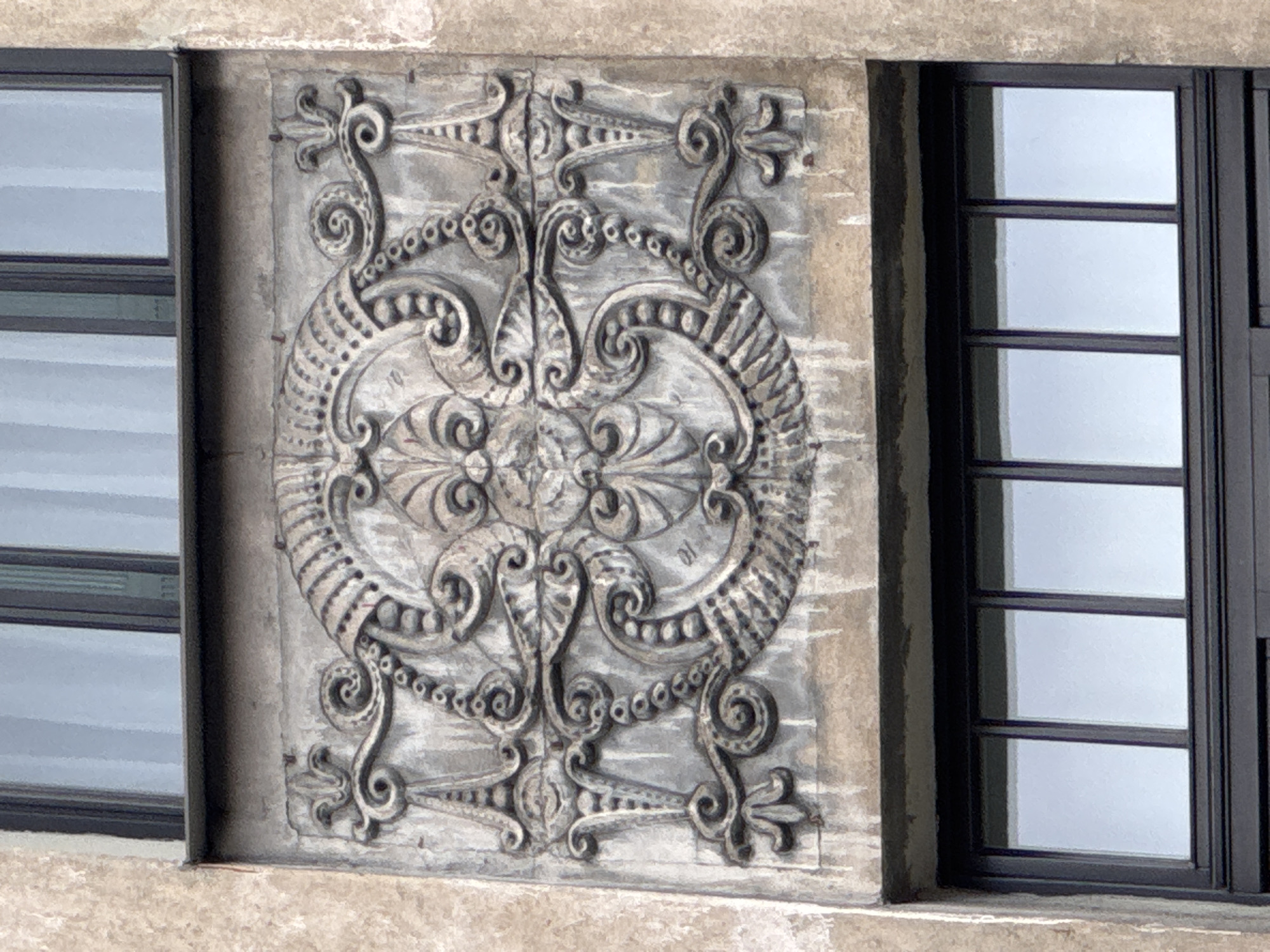 This is a close-up, eye-level outdoor shot of a decorative architectural bas-relief on the facade of a building in Riga, Latvia. The bas-relief is carved into a grey stone panel and features an ornate, symmetrical design with floral and scroll motifs. At the center is a stylized fleur-de-lis, surrounded by elaborate scrolling patterns, bead-like chains, and stylized leaves or petals. The relief shows signs of age and weathering, with some staining and pitting visible on the surface. Above and below the relief are dark-framed windows with multiple panes, reflecting a cloudy sky. The surrounding building material appears to be a textured, light-colored stucco or concrete. The image composition focuses entirely on the architectural detail, with no people or other discernible objects present. The specific location in Riga is not identifiable from the image itself.