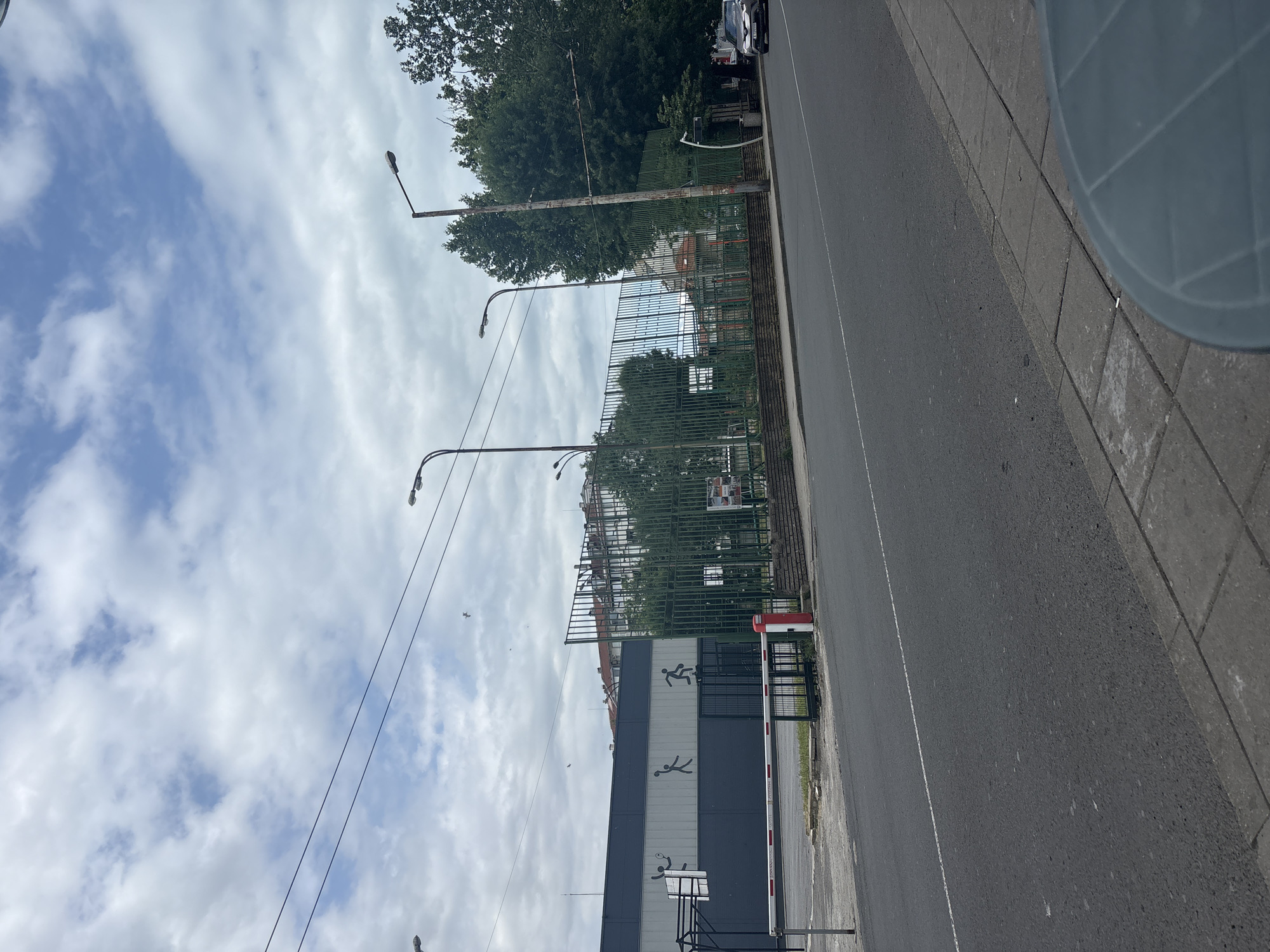 The image depicts a view of a street in the city of Burgas, Bulgaria, on a cloudy day. In the foreground, there is an asphalt road with a white line marking the lane. To the right of the road is a sidewalk made of concrete paving stones. Further back, a green metal fence encloses a sports court area. The court has a dark surface and is surrounded by trees and buildings. A white and red striped barrier is positioned at an entrance to the sports facility. The building behind the fence has a dark grey and white facade with silhouette graphics of people engaged in sports. Overhead, power lines are visible against a sky filled with white and grey clouds. There are also streetlights visible along the road. A car is parked on the right side of the road in the distance.