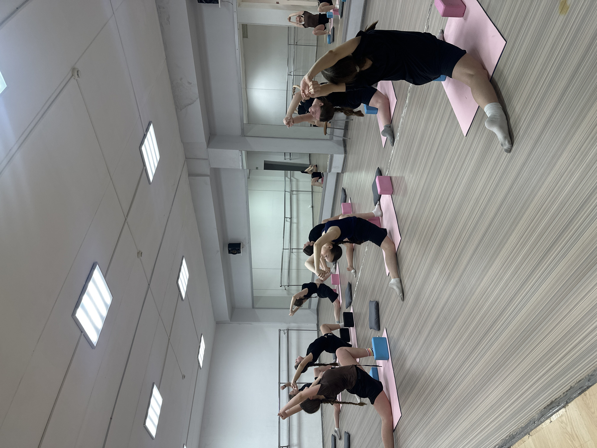 In an indoor studio located in Votkinsk, Russia, a group of individuals are engaged in a yoga session. They are performing a backbend pose on pink yoga mats. Some individuals are using yoga blocks. The studio has mirrors on the wall. The lighting is provided by square ceiling lights. The floor is a light wood color.