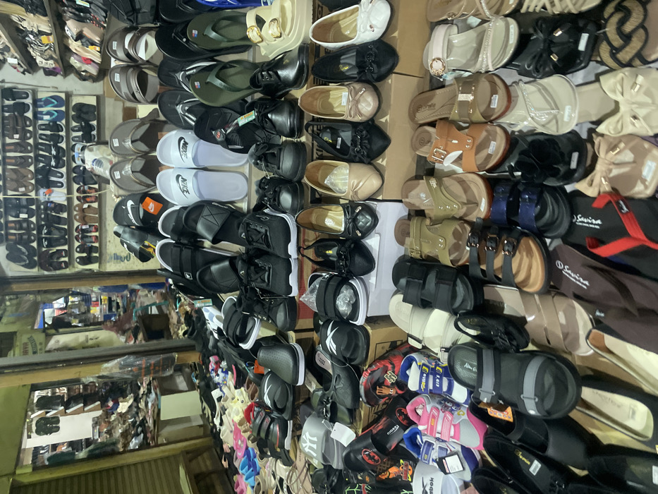 This image depicts a densely packed retail shoe display within a market or shop in Nganguk, Indonesia. Hundreds of footwear items are arranged on multiple tiered surfaces and shelves, extending from the foreground into the background. The selection includes a wide variety of styles such as athletic sneakers, casual slip-on shoes, dress flats with decorative bows, open-toed sandals, slide sandals (some bearing Nike branding), flip-flops, and children's shoes (some with Reebok branding). The footwear exhibits a diverse color palette including black, white, grey, beige, brown, pink, blue, red, and gold tones. Many items appear new, with some still displaying price tags or clear plastic packaging. In the background to the left, reflections on a vertical surface partially obscure additional merchandise and a rolled-down metal shutter. A blurry, partially obscured human figure, potentially wearing a headscarf, is faintly discernible in the distant left background. Wall-mounted shelves displaying more footwear are visible in the upper right quadrant, all illuminated by artificial lighting.