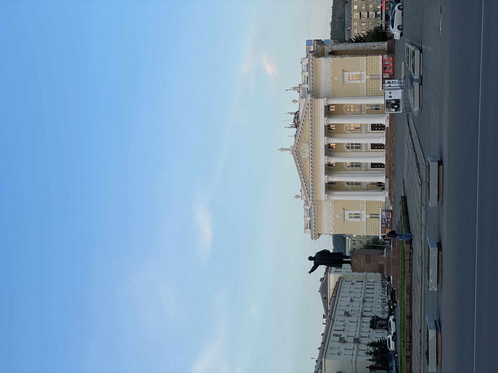 The image shows a view of a public square in Zheleznogorsk, Russia. A statue of Lenin stands in the foreground, pointing his right arm forward. Behind the statue is a large, pale yellow building with white columns, likely a public building or theater. The street is paved, with traffic present. Several people sit along the edge of the area, likely as relaxation. The sky is clear blue, with little cloud cover visible. Other buildings and trees can be seen in the background.