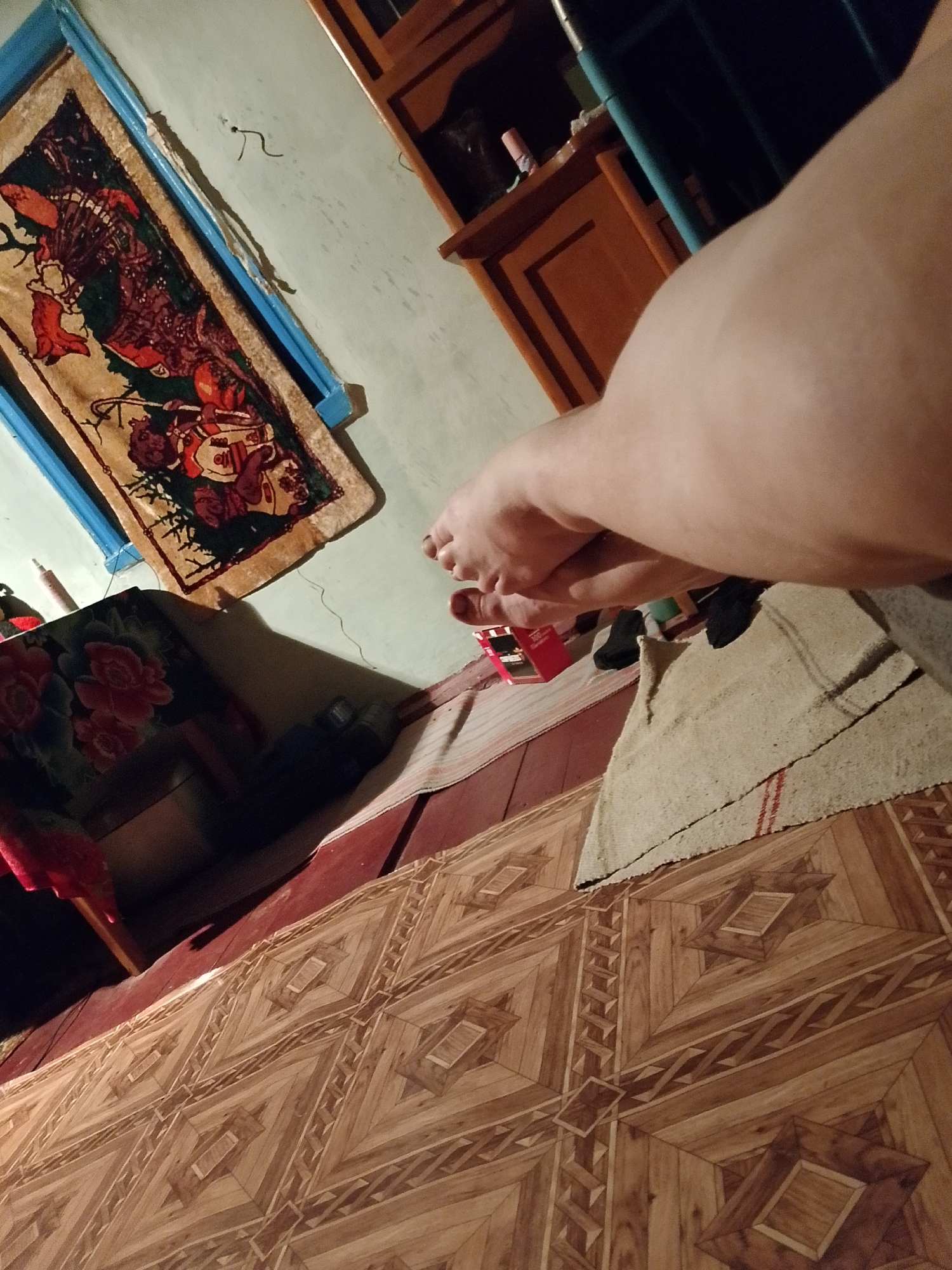Location: Unknown, Unknown.
This indoor, first-person perspective image captures a scene featuring two individuals, a domestic environment, and various objects. The camera's vantage point is low, looking upwards and across a patterned floor.
In the immediate foreground, to the right, a large, light-skinned human thigh/leg, belonging to the photographer, angles upwards and right, partially cropped. Below this limb, on the floor, is a small, rectangular rug made of light-colored, coarsely woven fabric with a single red stripe, laid over a reddish-brown floor surface.
In the midground, towards the center-left, are two light-skinned bare human feet, belonging to a second individual. The feet are angled slightly towards the photographer. To the immediate left of these feet is a red and white rectangular pack of cigarettes (e.g., Marlboro brand). Further back, behind the feet, two dark objects, likely shoes or socks, rest on the floor. Another small, light-colored, woven fabric rug lies on the floor to the left of the feet, extending diagonally towards the upper left.
The floor covering across the majority of the lower frame is a patterned laminate or linoleum designed to resemble parquet wood, featuring a repeating geometric design of squares, diamonds, and chevron patterns in various shades of brown wood grain. This patterned floor meets a plain reddish-brown floor surface beneath the rugs.
In the background, a light blue-green painted wall dominates the upper left. Mounted on this wall is a rectangular blue-painted wooden frame containing a detailed tapestry or wall hanging. The tapestry depicts stylized figures, possibly in traditional dress, in colors of red, dark green, and brown on a lighter beige background. Above the tapestry, a thin, dark, curved wire or hook is visible on the wall. Below the tapestry, a dark wooden table or cabinet is present, partially covered by a dark cloth with a red floral pattern. A light-colored cylindrical object, possibly a vape pen or cosmetic tube, stands on this table. To the upper right background, a dark brown wooden cabinet or shelving unit is partially visible, obscured by the photographer's leg.
The scene suggests a relaxed, informal setting, possibly a living area, with both individuals situated at floor level.