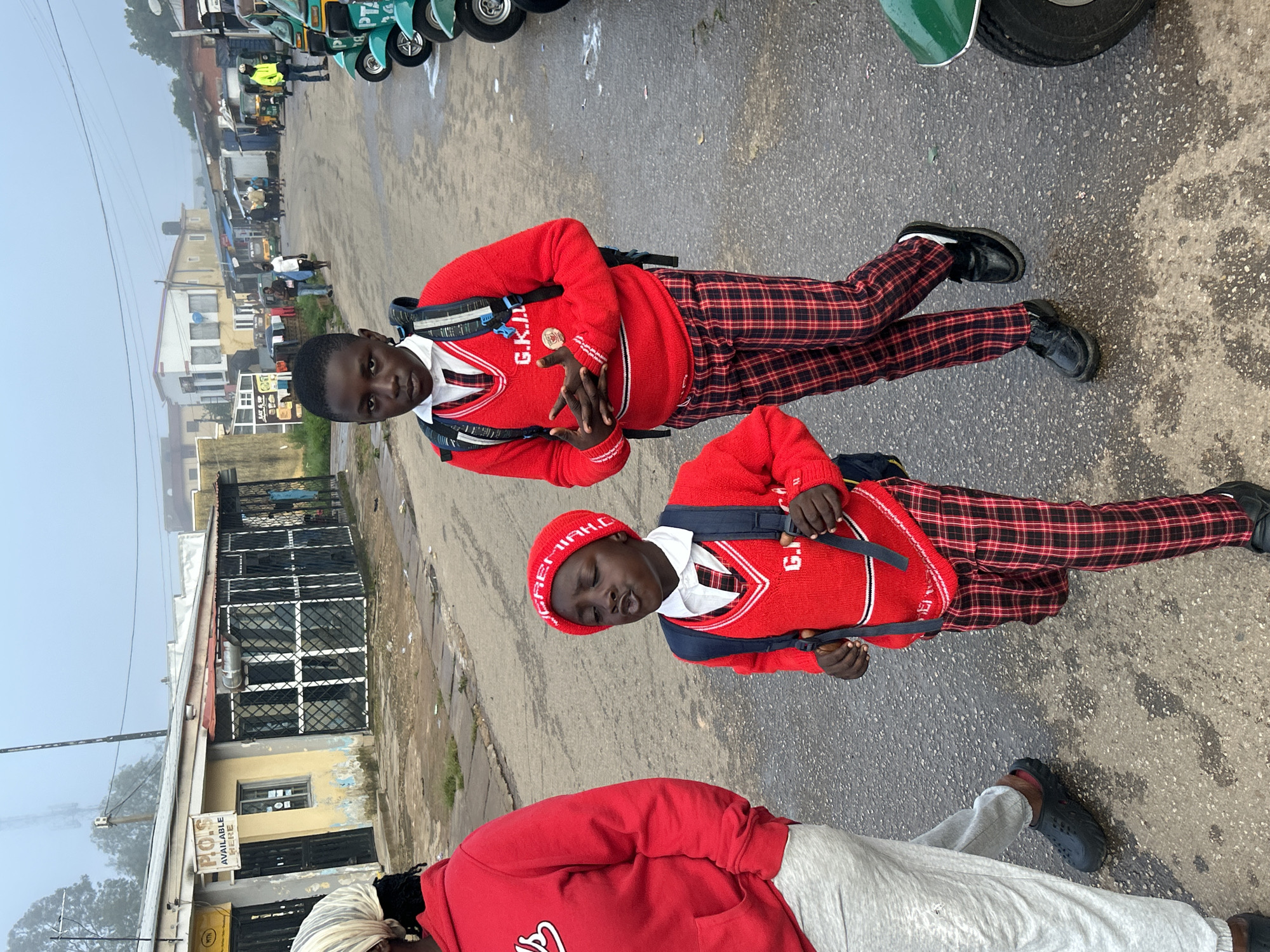 The image depicts a street scene in Jos, Nigeria, likely during a foggy morning given the atmospheric haze. In the foreground, two children are walking on a wet asphalt road. The child on the right, wearing a red sweater vest over a white shirt and tie, red and black plaid pants, black shoes, and a black backpack, is looking towards the camera with a serious expression. The child on the left is partially visible, wearing a red hooded sweatshirt and light grey sweatpants. Behind them, other people and various buildings line the street, with a row of green and white tricycles parked on the right side of the road. One building in the background features a sign indicating "P.O.S. AVAILABLE HERE." The overall environment suggests a commercial area with some light industrial or residential structures.