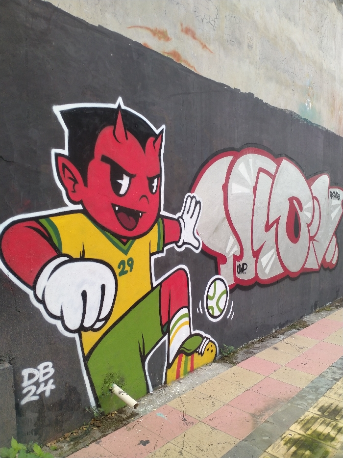 This media file features a vibrant graffiti mural painted on a textured concrete wall in Karanganyar, Indonesia. The central subject is a cartoon character, depicted as a red-skinned, black-haired boy with small horns, pointed ears, and sharp fangs, giving him an impish or devil-like appearance.
The character is actively engaged in what appears to be a soccer game. He is shown mid-action, seemingly kicking a small, stylized soccer ball with green markings. He wears a yellow soccer jersey with green accents, emblazoned with the number "29" on the front. His attire also includes green shorts, and distinctive red, yellow, and white striped socks paired with yellow shoes. His expression is energetic and somewhat mischievous, with wide eyes and a visible smile revealing his fangs.
To the right of the character, the word "SLORK" is painted in a large, bubble-style graffiti font, outlined in red with a light grey fill that has a radiant line pattern. Below "SLORK," a smaller tag reads "UMP," and another tag, "KRESNA," is visible above and to the right. On the lower left section of the wall, near the character's feet, the tag "DB 24" is present.
A notable detail is the clever integration of a real-world object into the artwork: a white PVC pipe protrudes from the base of the wall, appearing to emerge from beneath the character's green shorts. The wall itself is dark grey in its lower section, transitioning to a lighter, weathered grey above. The scene is set outdoors, with a paved sidewalk featuring pink, yellow, and grey tiles visible in the foreground. The lighting suggests it is daytime.