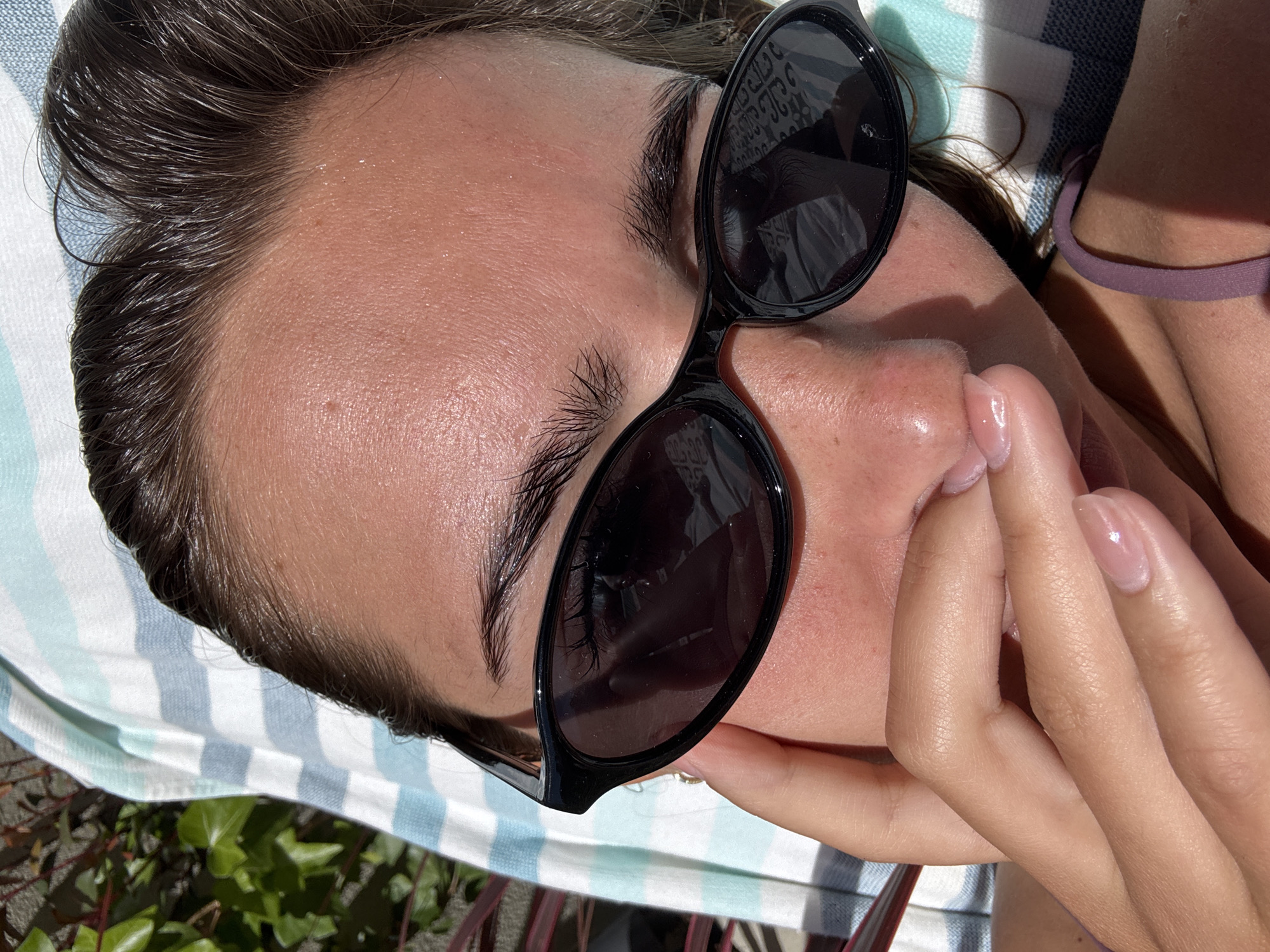 A close-up, high-angle photograph shows a person with light skin lying on a striped blue and white towel outdoors. The person is wearing sunglasses with dark lenses and black frames, and their dark hair is slicked back. The subject's hand is near their face. The background includes green foliage. There is no definitive information in the image that identifies the location as Porto, Portugal.