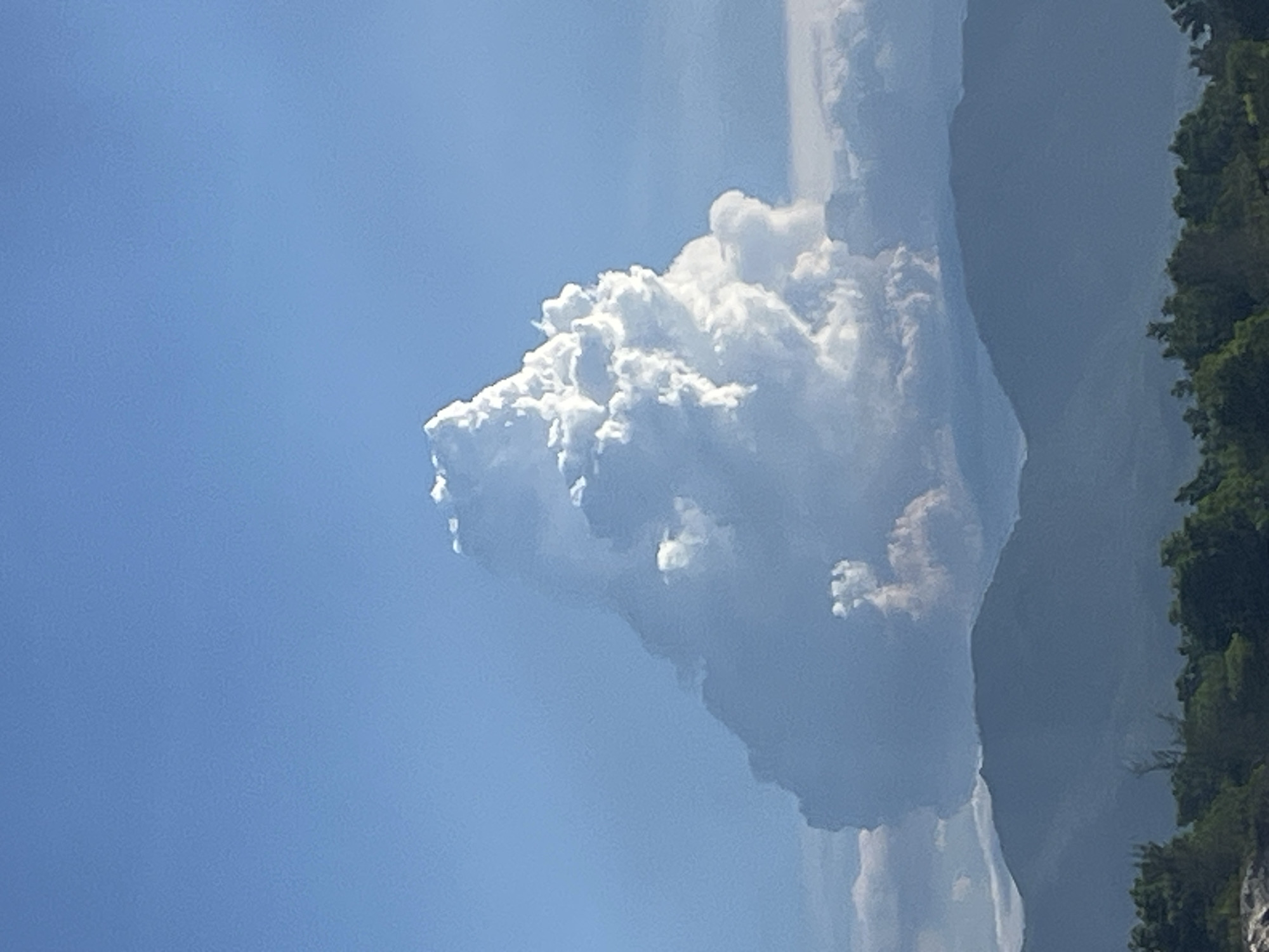 A towering cumulus cloud, resembling a mountain, dominates the clear blue sky. The cloud is brightly illuminated from above, with its base appearing darker and tinged with pink. Below the cloud, layers of hazy blue mountains recede into the distance. The foreground is filled with a dense line of dark green trees. The scene is set in Bac Cam Ranh Ward, Vietnam.