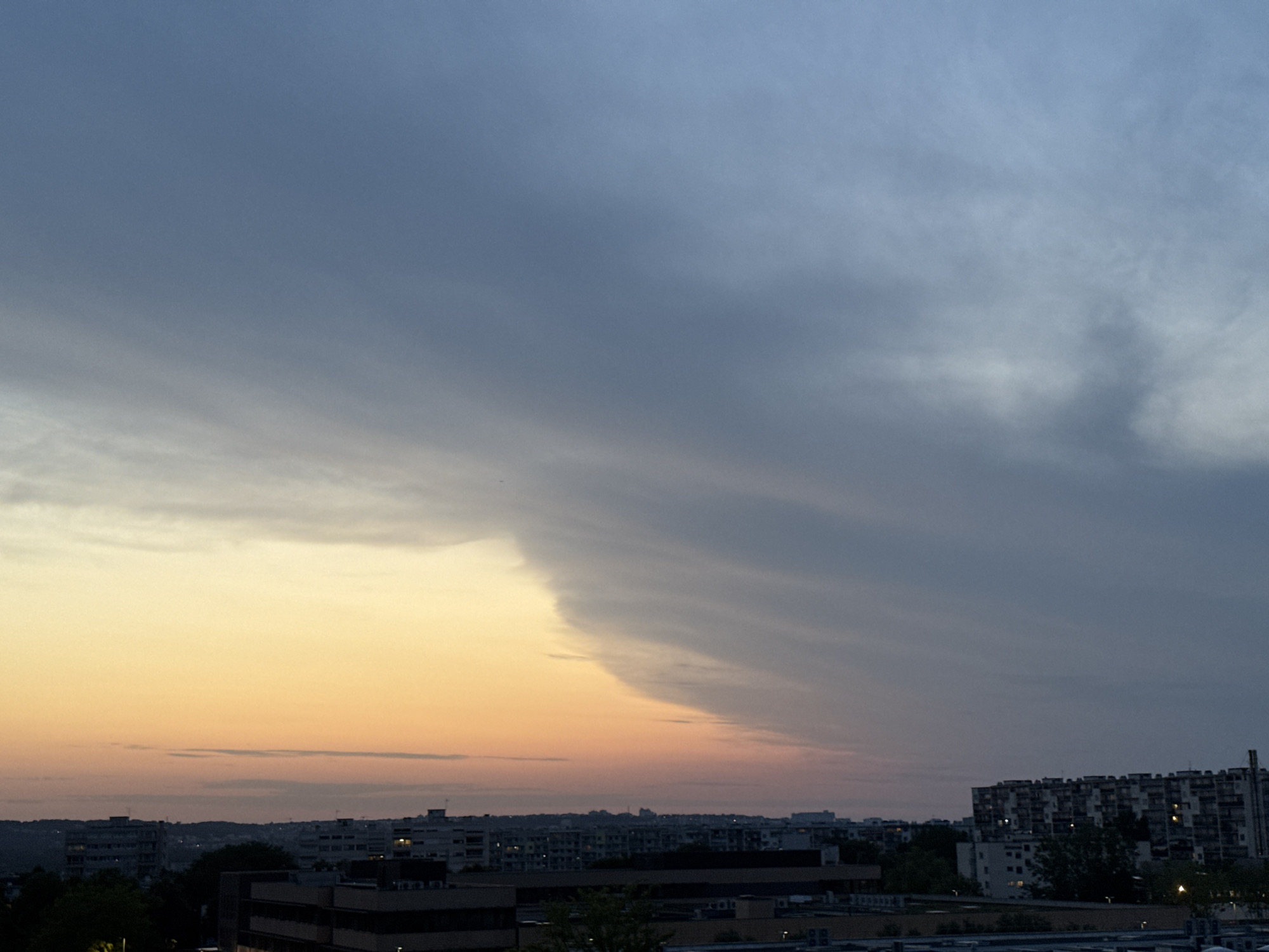 This is an outdoor, high-angle medium shot of a cityscape at dusk. The sky dominates the upper two-thirds of the frame and is a gradient of colors transitioning from a pale orange-yellow on the left to a deep blue-gray on the right. Wispy clouds streak across the sky, creating a sense of depth and movement. A sharp line divides the lighter sky from the darker sky, suggesting the edge of a weather front.
Below the sky, the city skyline is visible. A dense collection of buildings stretches across the horizon. On the right, a large, multi-story building with many windows stands out. To its left, other buildings of varying heights are visible, some appearing as silhouettes against the twilight. Trees and darker building structures are in the foreground, indicating that the photo was taken from a higher vantage point, likely another building. The overall impression is one of a city transitioning from day to night under a dramatic sky. There are no people or specific actions depicted in the scene.