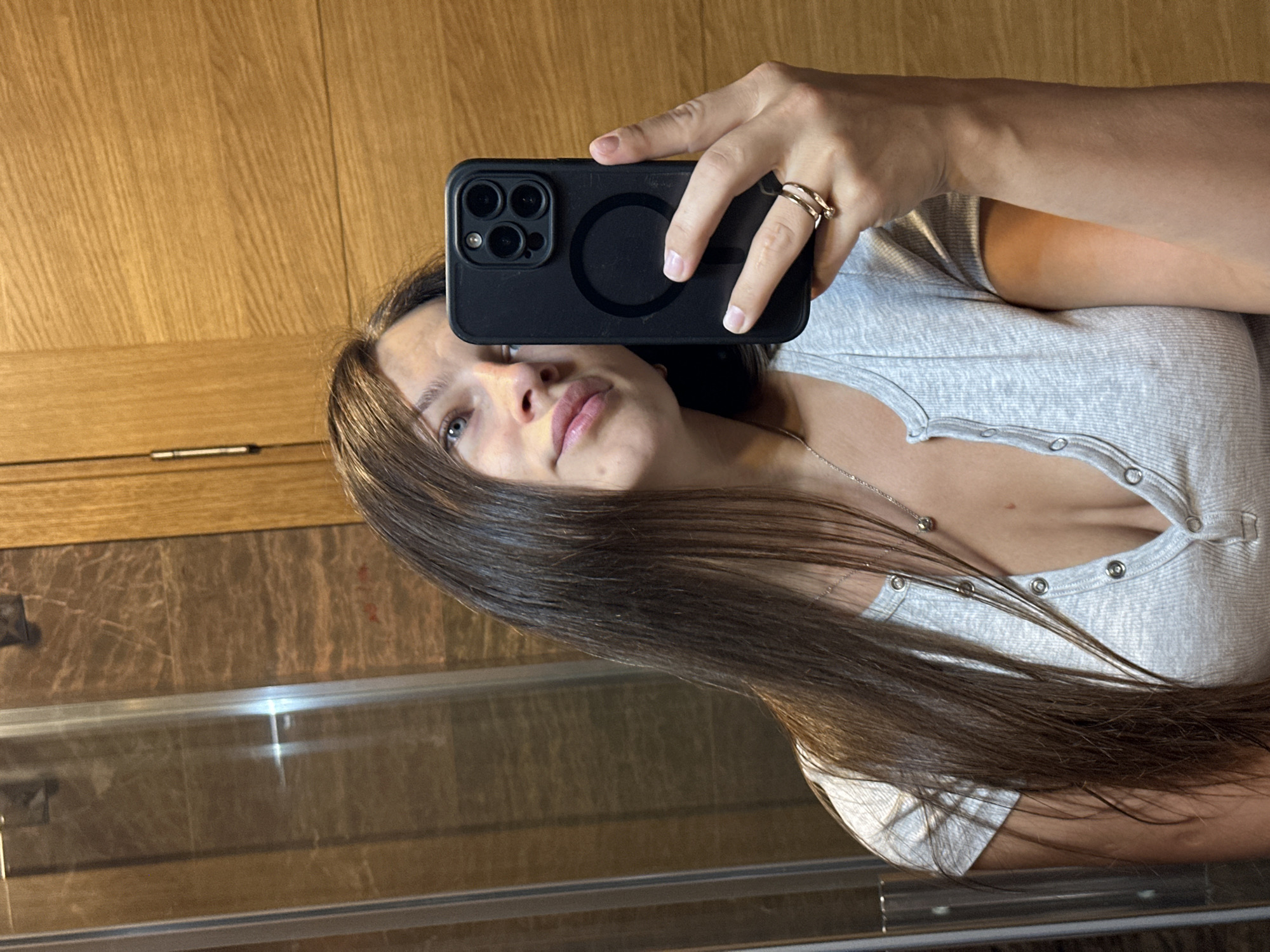 A young woman with long brown hair and blue eyes is taking a selfie in a bathroom. She is wearing a grey ribbed Henley shirt and a silver necklace. She is also wearing two rings on her right hand, which is holding a black iPhone in a case. The background appears to be a bathroom with wood-paneled walls and a glass shower door. The specific location is a bathroom in Donji Pobori, Montenegro.