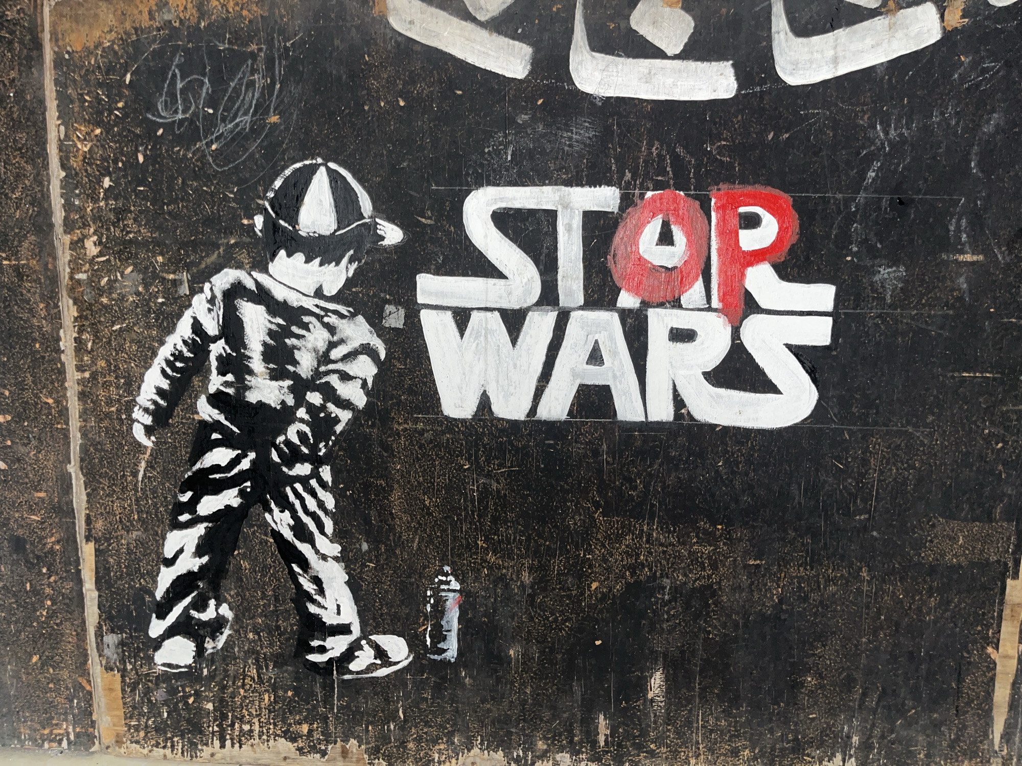 This media file shows a close-up of graffiti on a dark, weathered surface, likely a wall or wooden panel. The artwork depicts a stencil of a child wearing a striped outfit and a baseball cap, walking towards the right side of the frame. To the right of the child, large white letters spell out "STOP WARS," with the "OP" in "STOP" colored in red. A miniature spray paint can, also rendered in stencil style, is visible near the child's feet. The background is black with visible textures and scratches, suggesting an urban or outdoor environment. The overall impression is one of protest or social commentary, using the popular "Star Wars" franchise title as a message against conflict. The style is reminiscent of street art, with bold lines and contrasting colors. The context of Ubud District, Indonesia, adds a layer of potential cultural interpretation to the artwork.