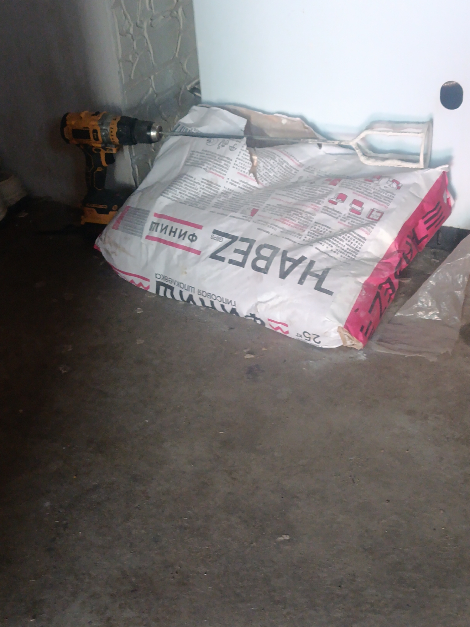 In an indoor environment with a concrete floor, a yellow and black cordless drill with a chuck is positioned on the left side of the frame, leaning against a textured wall. Next to the drill, a large, white bag of construction material, likely plaster or grout, is prominent. The bag features bold text that reads "HABEZ" and "МИНИФ" in Cyrillic. A mixing paddle, coated with white residue, rests on top of the bag. The background includes a plain white wall with a small, circular hole. The scene suggests a construction or renovation area.