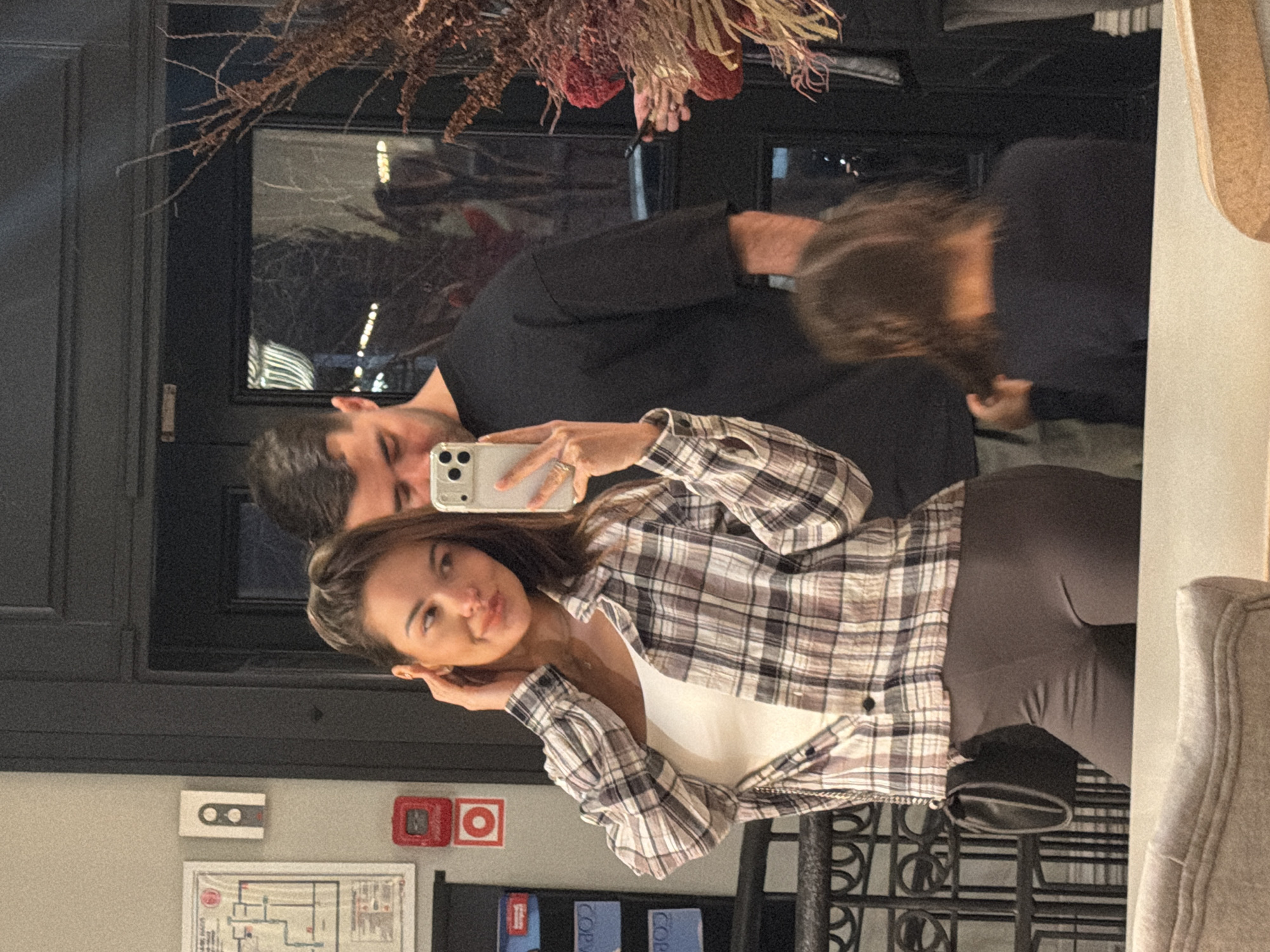 Three individuals are reflected in a mirror. The woman in the foreground is holding a phone to take a selfie. She is wearing a plaid shirt and leggings. Beside her is a man in a black shirt, and partially visible behind them is another person. The location appears to be indoors, possibly in a restaurant based on the decor. The absence of specific, clear location markers prevents the identification of a specific site in Krasnoyarsk, Russia.