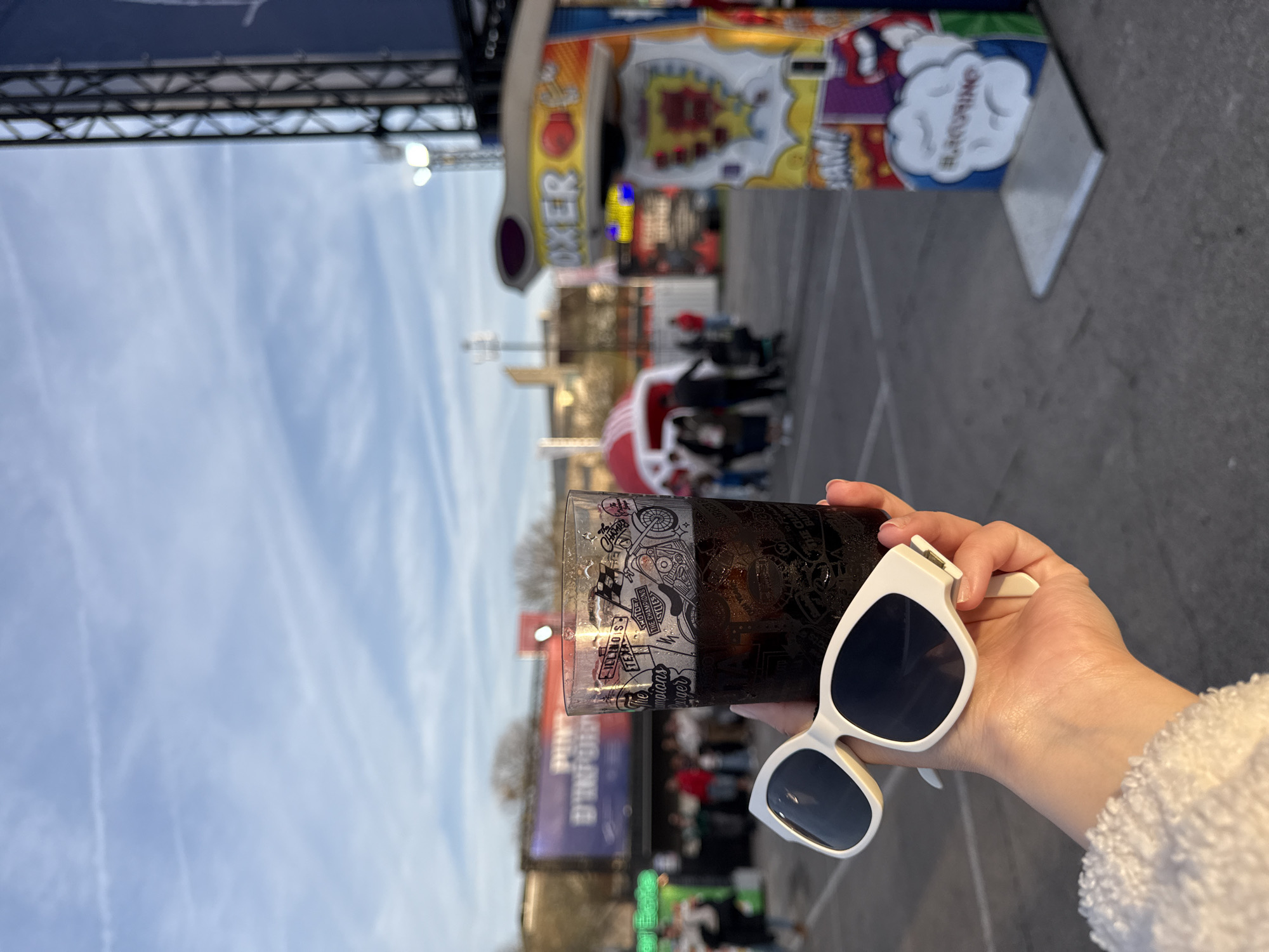 Here is a detailed description of the image:
**Scene:**
The image is taken at an amusement park, which is very likely located at La Fira de Montjuïc in Barcelona, Spain, as implied by the text "d'Import" seen on the poster in the background. The shot is an upward angle, suggesting the photograph was taken from a ground-level perspective. The picture is somewhat distorted due to the camera angle.
**Subjects & Objects:**
The main focus is a glass of what appears to be a dark liquid (possibly cola or a dark beer) and a pair of white sunglasses held in a person's hand. The glass has an elaborate design of motorcycles and racing motifs. In the background, there are various amusement park attractions including:
* A "Boxer" game stall
* An "El Chupete" sign
* Various banners and signs, some of which appear to have the words "d'Import".
* A crowd of people milling around the amusement park
**Environment:**
The environment includes a fairground or amusement park with various attractions. The day appears to be late afternoon or early evening as indicated by the position of the sun and the lighting. The sky is partly cloudy.
**People & Interactions:**
There is a crowd of people in the background, presumably visitors to the amusement park. However, there is no clear interaction, though the presence of the drink and sunglasses being held suggests someone is present and enjoying themselves.
