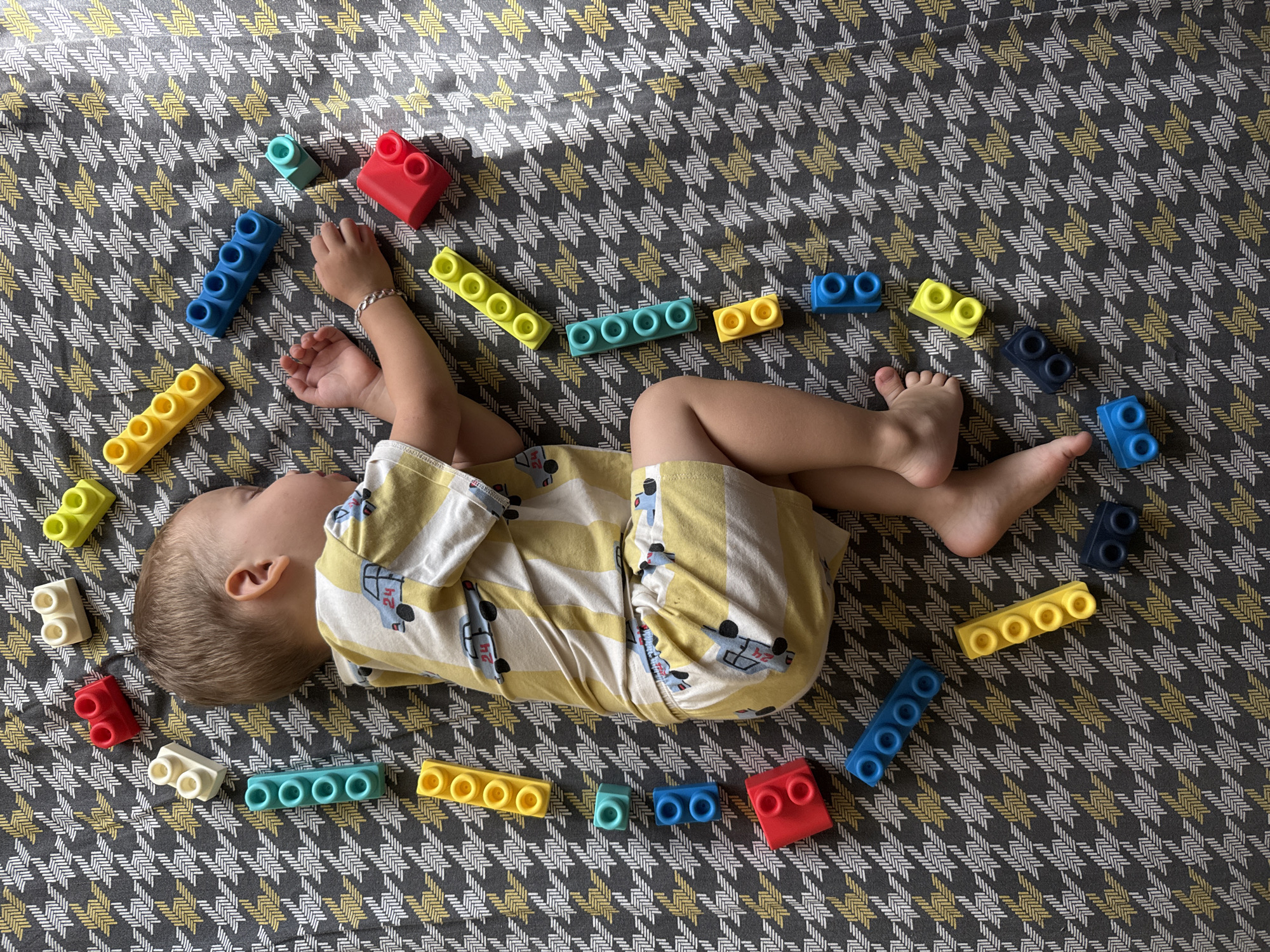 A young child is sleeping on their side on a patterned fabric surface. The child is wearing a yellow and white striped shirt with a car print. Around the child, colorful building blocks are scattered in a circular pattern. The blocks are red, yellow, blue, teal, and white. The fabric has a houndstooth-like pattern with alternating grey, white, and yellow lines. There is no indication of the location being Rusanovo, Russia, or any specific city.