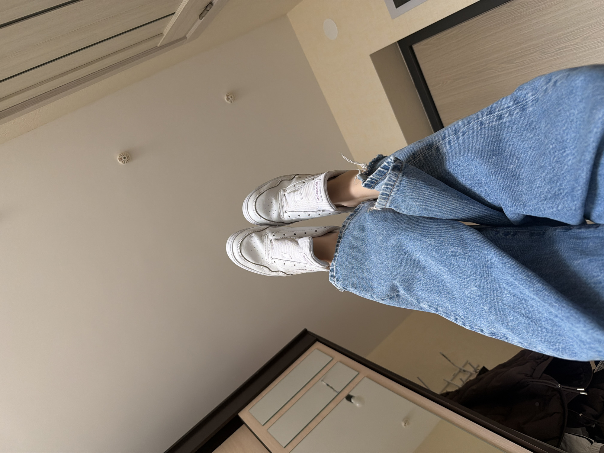 The image shows a person's legs and feet in a seemingly indoor setting. The person is wearing light blue jeans with frayed hems and white sneakers, which appear to be Reebok brand. The background shows a ceiling, a wall, a closet, and a mirror. There is a smoke detector on the ceiling. This image may have been taken in a residential space. There is no evidence to connect this image to Izhevsk, Russia.