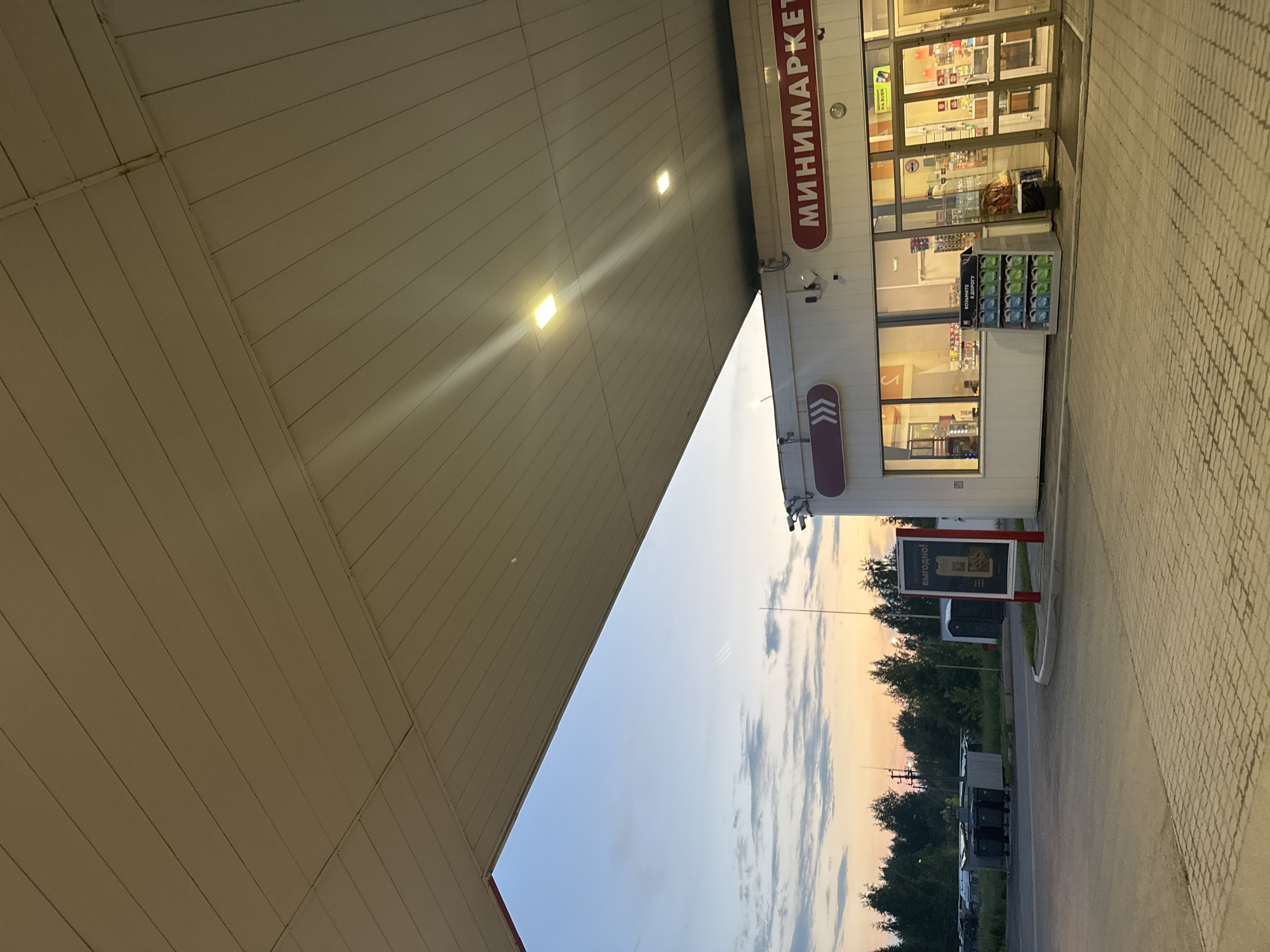 The image depicts the exterior of a minimarket located at a gas station, with a portion of the canopy and the sky visible. The minimarket has large windows and glass doors, with signs displaying "МИНИМАРКЕТ" in red lettering. Inside, shelves stocked with products are visible, and a display of cleaning supplies is positioned outside the entrance. In the background, trees and a cloudy sky at dusk or dawn are present. The ground is paved with interlocking bricks. The presence of trash bins, a portable toilet, and a sign with Cyrillic text suggest a location in Russia, likely in Krasnokamsk given the prompt. No people are clearly identifiable in the scene, though some indistinct figures may be present behind the glass windows. The scene is static and appears to be captured during twilight hours.