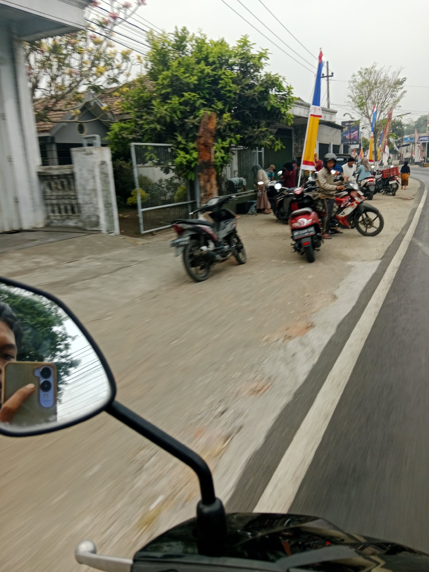 In Ringinrejo, Indonesia, on a cloudy day, a motorcycle rider captures a reflection in their side mirror showing themselves holding a smartphone. The rider is on a street lined with houses and trees. Several motorcycles are parked or being ridden on the side of the road. Flags are displayed along the street, indicating a possible local event or celebration. The road has a light gray asphalt surface with white lane markings. The scene appears to be a typical street in a residential or commercial area of Ringinrejo.