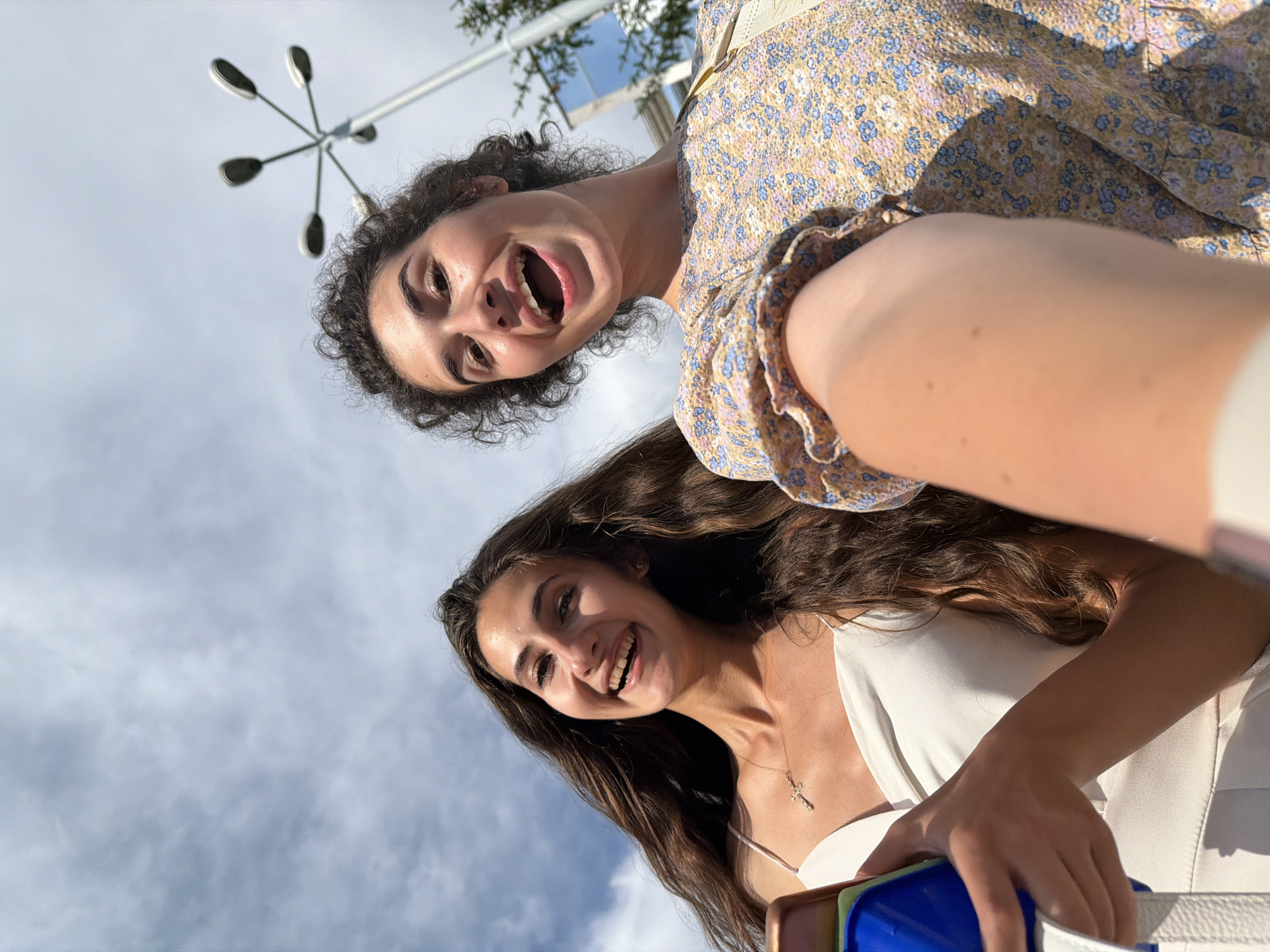 Two young women are captured in a low-angle, upward shot, looking into the camera with wide smiles. The woman in the foreground, on the right side of the frame, has long, dark, wavy hair and is wearing a white top with a delicate cross necklace. She is laughing, her eyes bright and her mouth open. Behind her and slightly to the left, another woman with curly dark hair is also smiling broadly, her mouth open in a joyful expression. She is wearing a floral-print dress with a yellow base and blue and pink flowers.
The background is a bright sky with scattered clouds, suggesting it is daytime. A street lamp with multiple lights extends from the upper left of the frame, indicating an outdoor urban or public setting. The lighting suggests it is a sunny day, casting shadows and highlighting the subjects' features. The image conveys a sense of happiness and shared enjoyment between the two individuals. The camera angle creates a dynamic and engaging perspective, making the viewer feel as though they are looking up at the women from below.