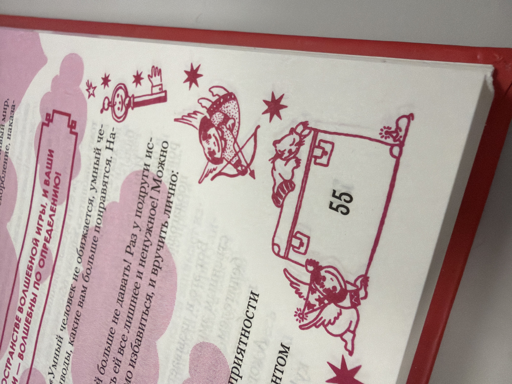 The image shows a close-up of an open book. The pages are white with pink text and illustrations. The text appears to be in Russian and includes phrases like "ОСТРАНСТВО ВОЛШЕБНОЙ ИГРЫ" and "УМНЫЙ ЧЕЛОВЕК". The illustrations are in red and feature cartoonish figures, including a key with a face, a cupid with a bow and arrow, and a treasure chest with a number "55" on it. The binding of the book is red. The background is a plain light gray surface. There is no indication of the city of Homyel, Belarus in the image itself.
