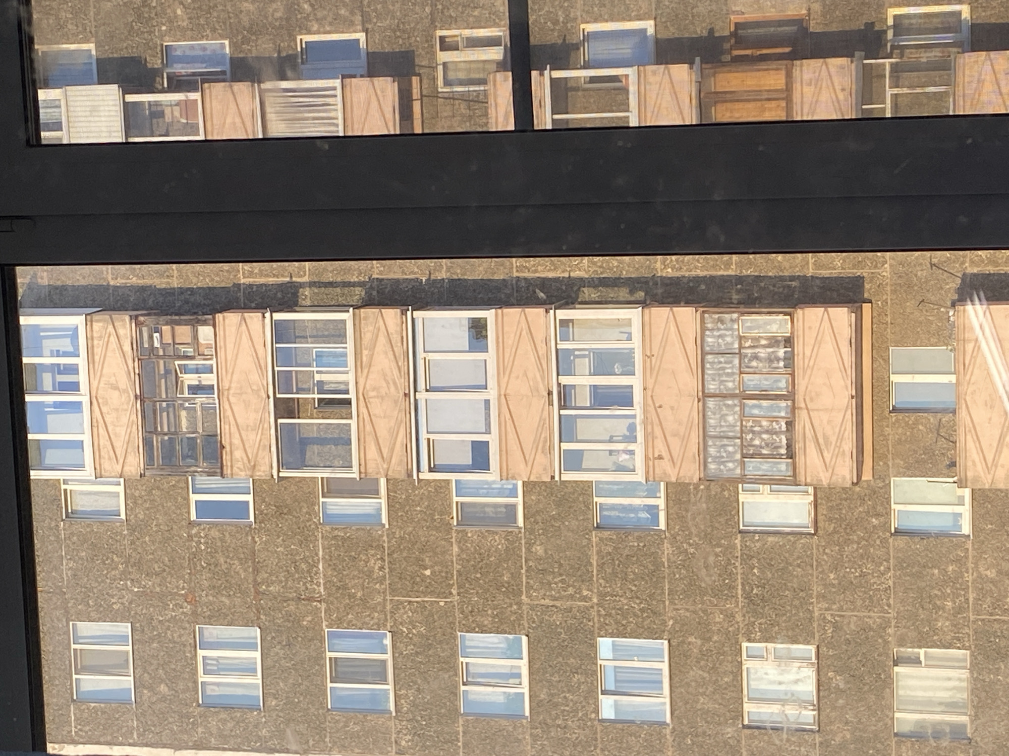The image depicts the exterior of an apartment building in Votkinsk, Russia, as seen from an elevated perspective through a window. The building is characterized by a concrete facade with a repetitive pattern of windows. Many of the windows have shutters, some of which are open and some closed. The upper portion of the frame shows the reflections of similar buildings across the street.
