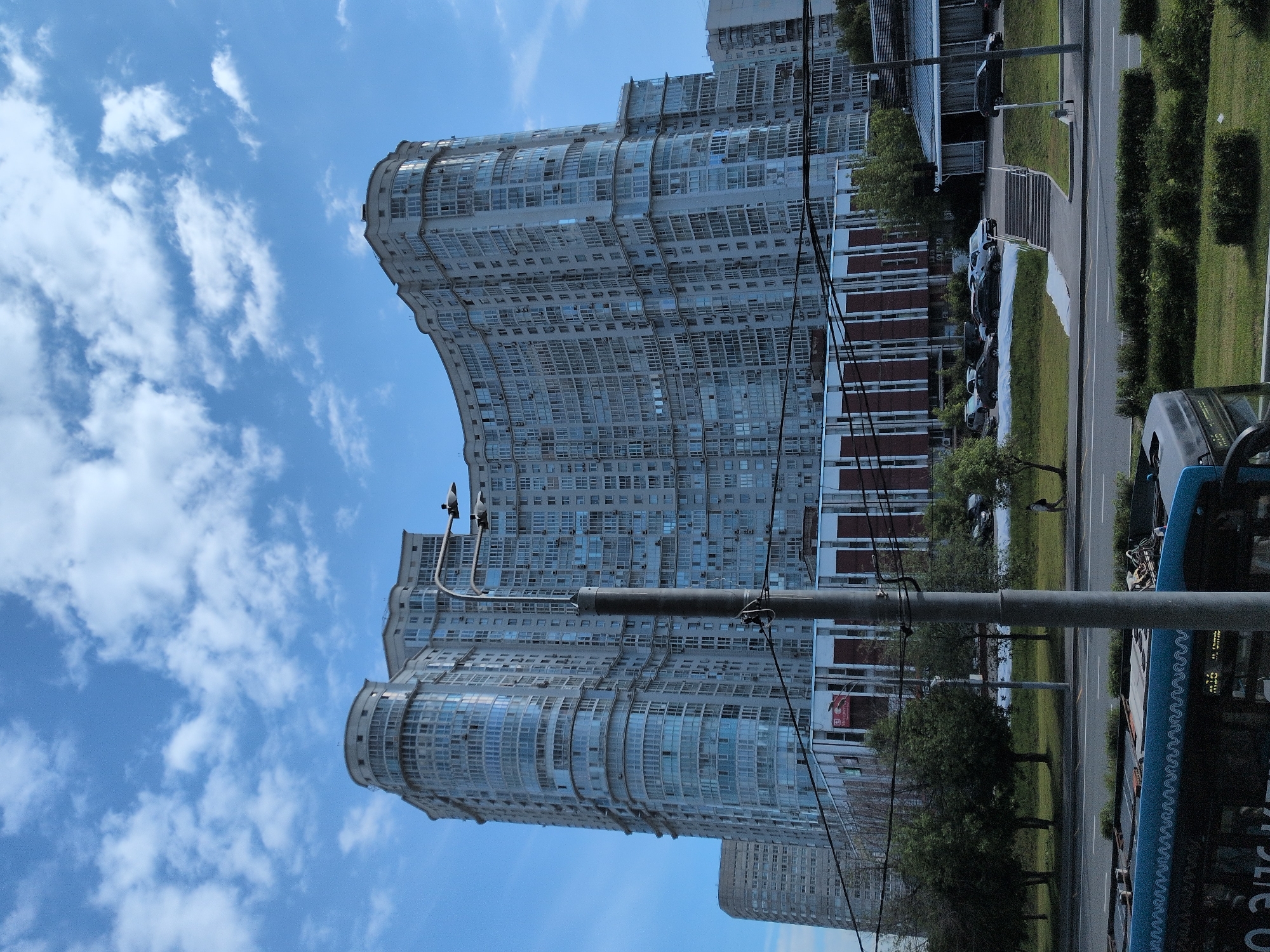 The image depicts a view of residential buildings in Moscow, Russia. The architecture includes curved facades with numerous windows, indicating a modern residential complex. A street runs alongside the buildings, with parked cars and a visible bus. There are trees and greenery along the roadside. Overhead utility lines crisscross the scene. The sky is blue with scattered clouds. The image appears to be taken from a low angle, close to ground level.
