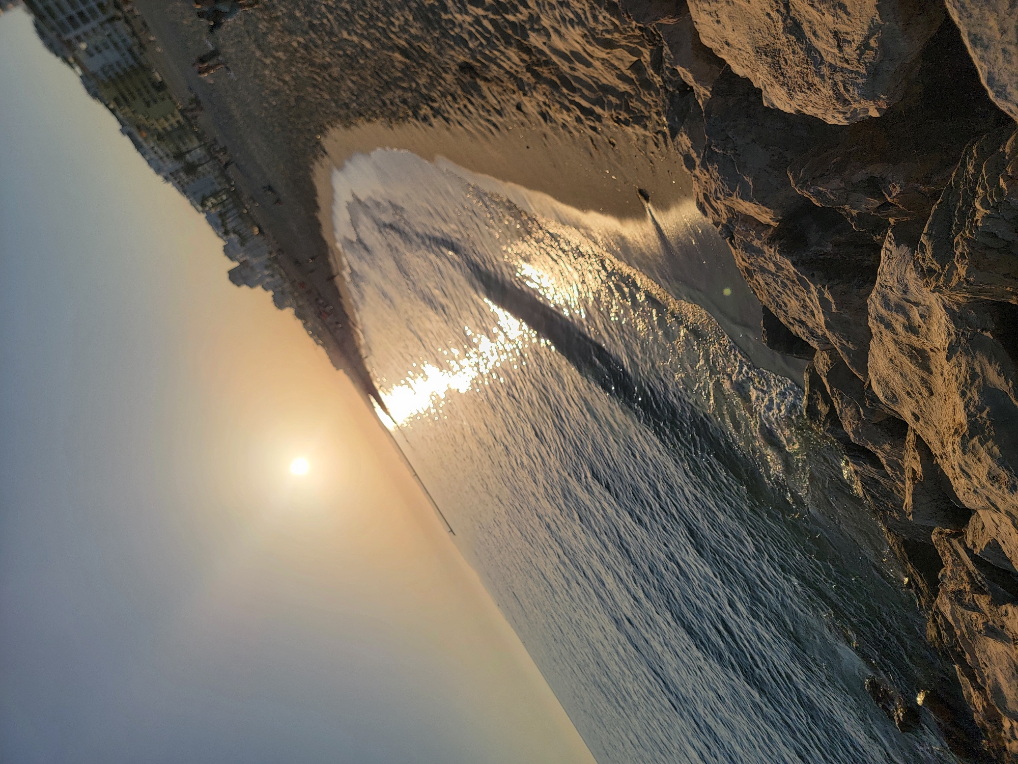 This image depicts a serene beach scene at Quarteira, Portugal, during what appears to be sunset or sunrise. The sun, a bright orb low in the sky, casts a warm, golden glow across the water and sand, creating a shimmering effect on the gentle waves as they meet the shore.
The foreground is dominated by a rocky shoreline on the right, with large, rough-textured rocks catching the sunlight and casting shadows. To the left, the calm sea stretches towards a sandy beach. The wet sand near the water's edge reflects the sky and the sunlight. Further down the beach, a line of buildings, likely hotels or apartments, stands parallel to the coastline, suggesting a developed resort area. A few indistinct figures can be seen on the beach and along the promenade in front of the buildings.
The overall mood is tranquil and peaceful, evoked by the soft light, the gentle motion of the waves, and the vastness of the sky. The muted colors of the sky and the warm tones of the sunlight contribute to a sense of calm and natural beauty. The scene captures the quiet moments of a day at the beach as the light begins to fade or emerge. There is no visible text in the image.